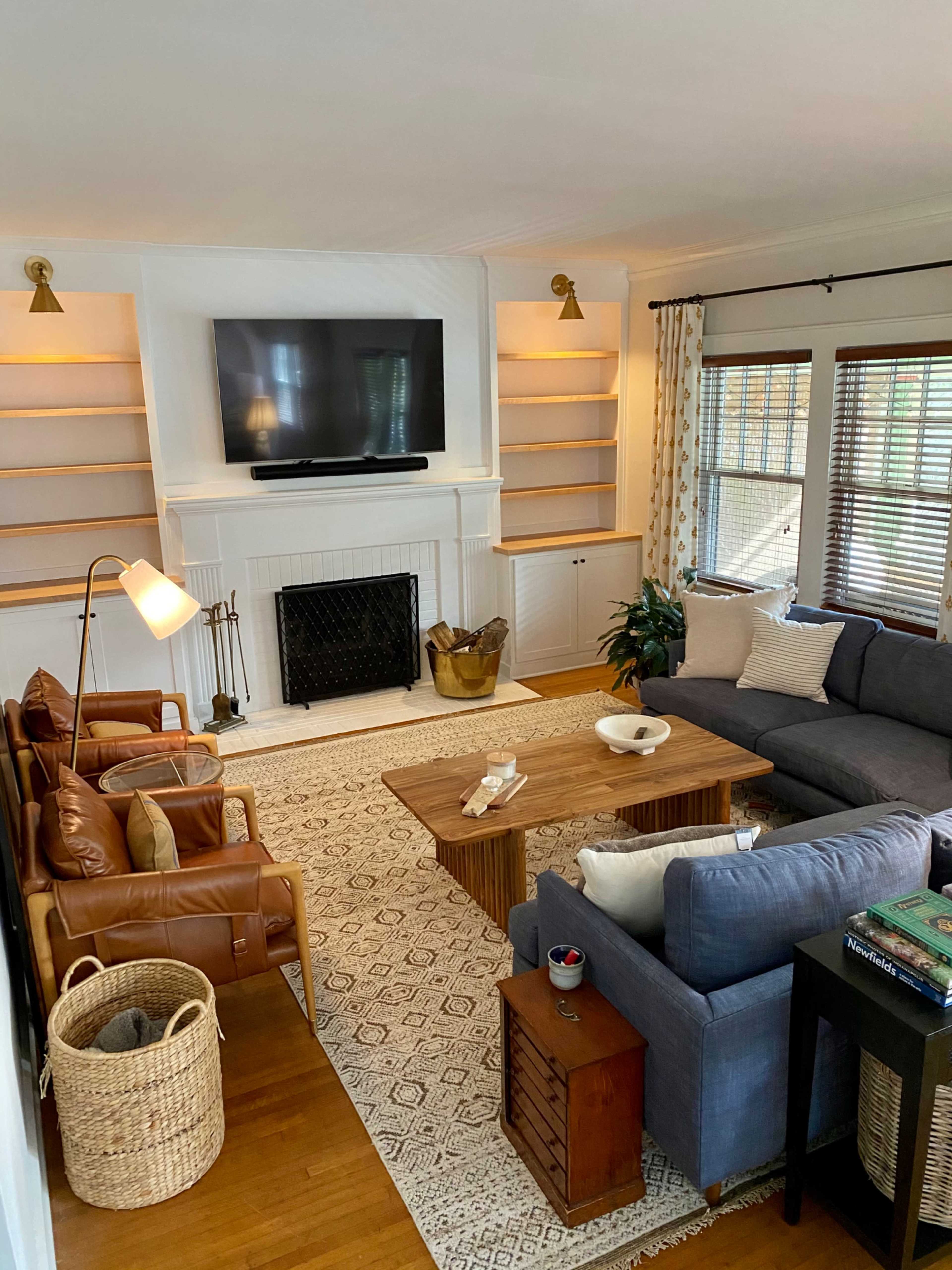 The living room features a seating arrangement with a brown leather chair, a blue sofa, a wooden coffee table, and shelves beside a fireplace beneath a mounted television.