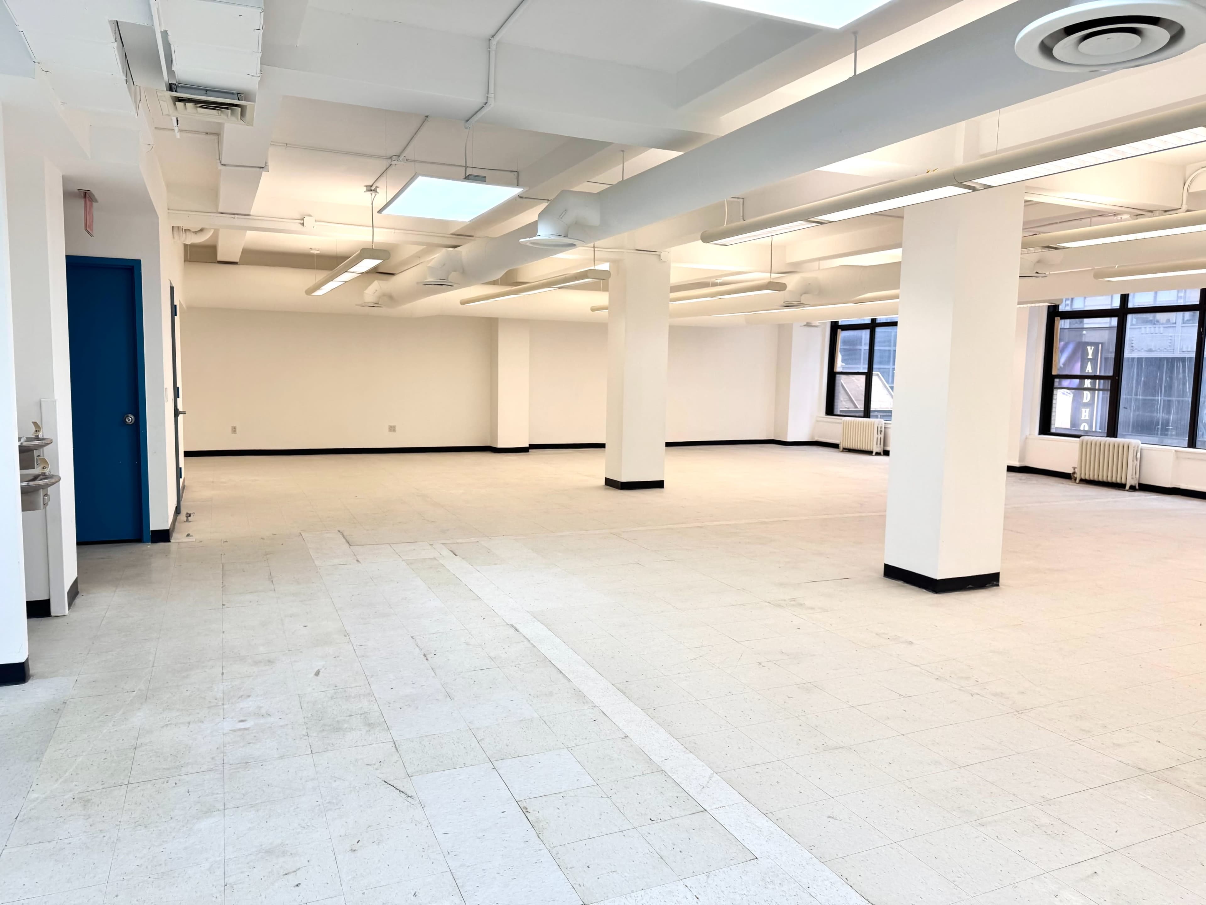 The image shows a vast, empty office space with white walls, multiple ceiling lights, and large windows.