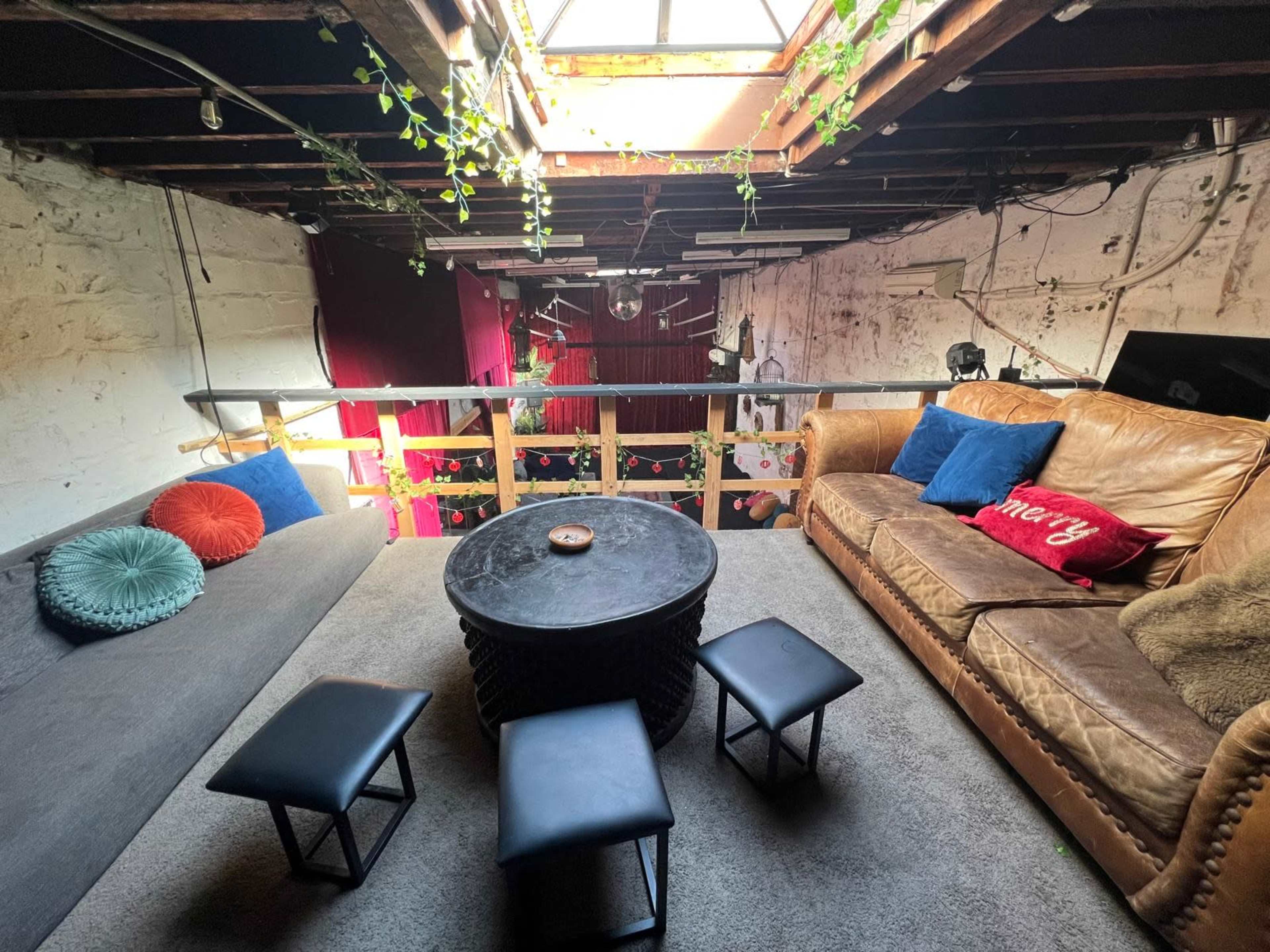 The image shows a cozy loft living area with a sofa, several small stools, and a circular coffee table under a skylight, surrounded by decorative plants.