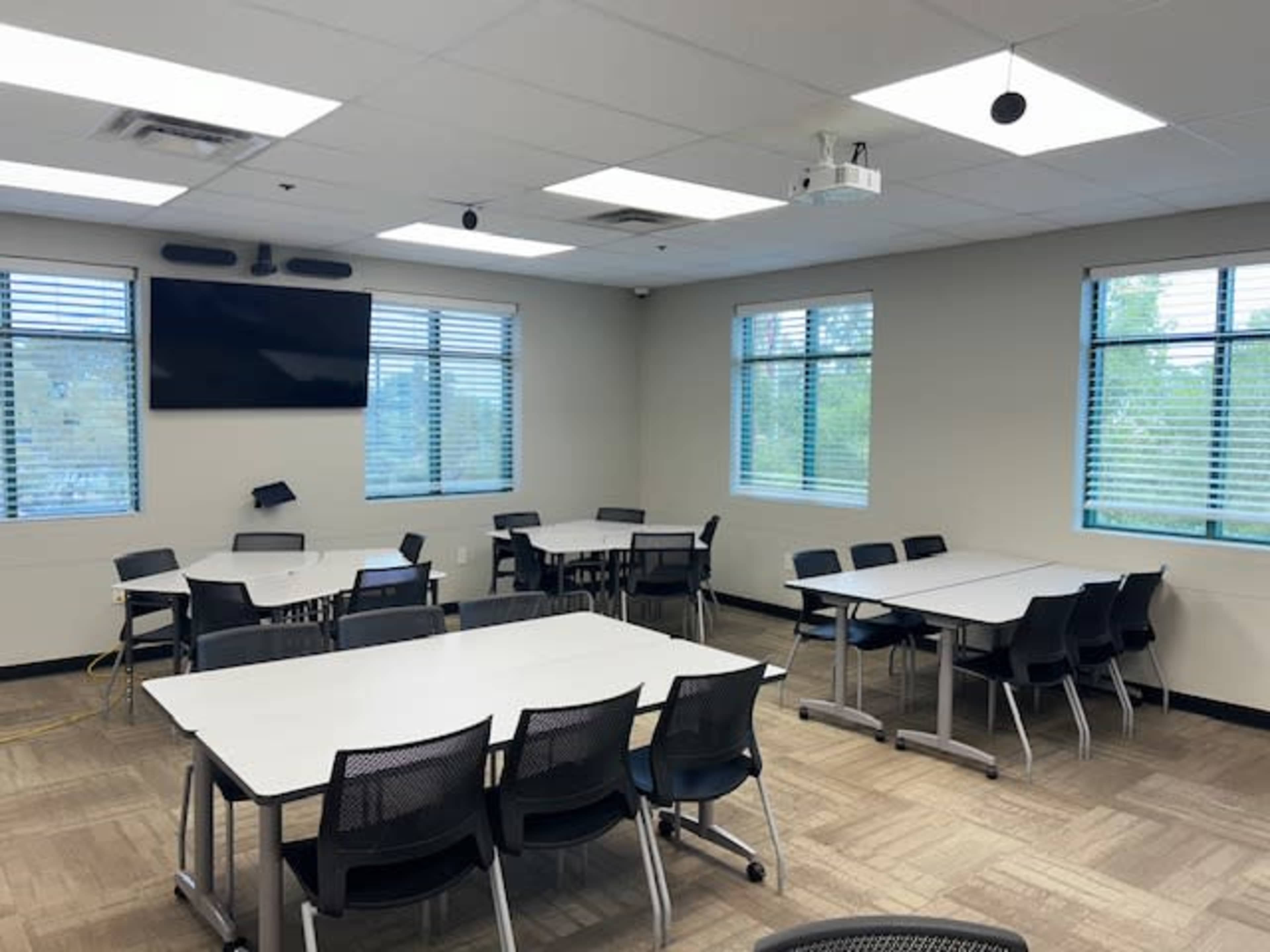 A well-lit classroom features multiple tables and chairs arranged for meetings or group work, with a projector and screen mounted on the wall.