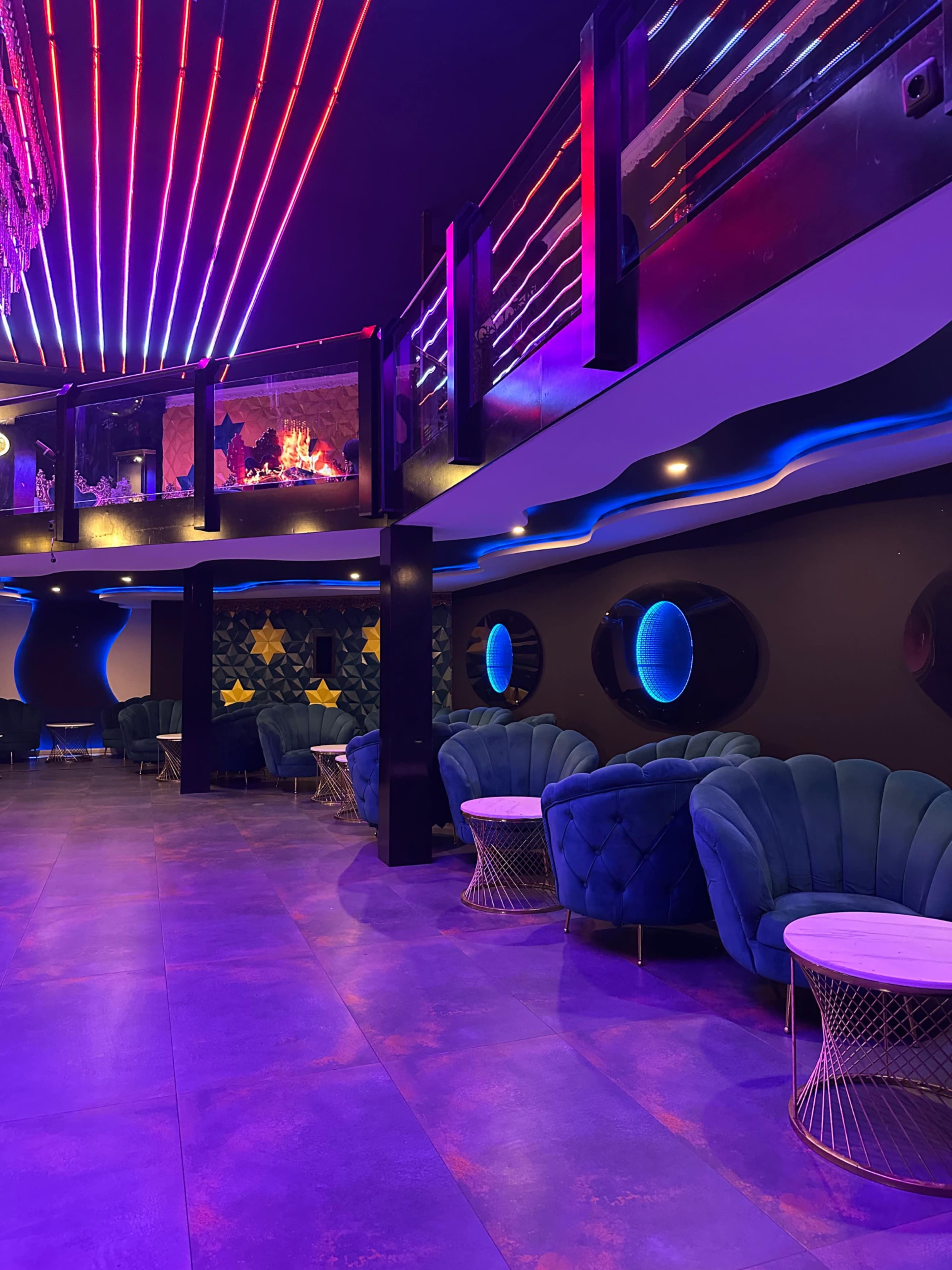 The image shows a modern lounge with dark walls, plush blue seating, and colorful neon lighting overhead.