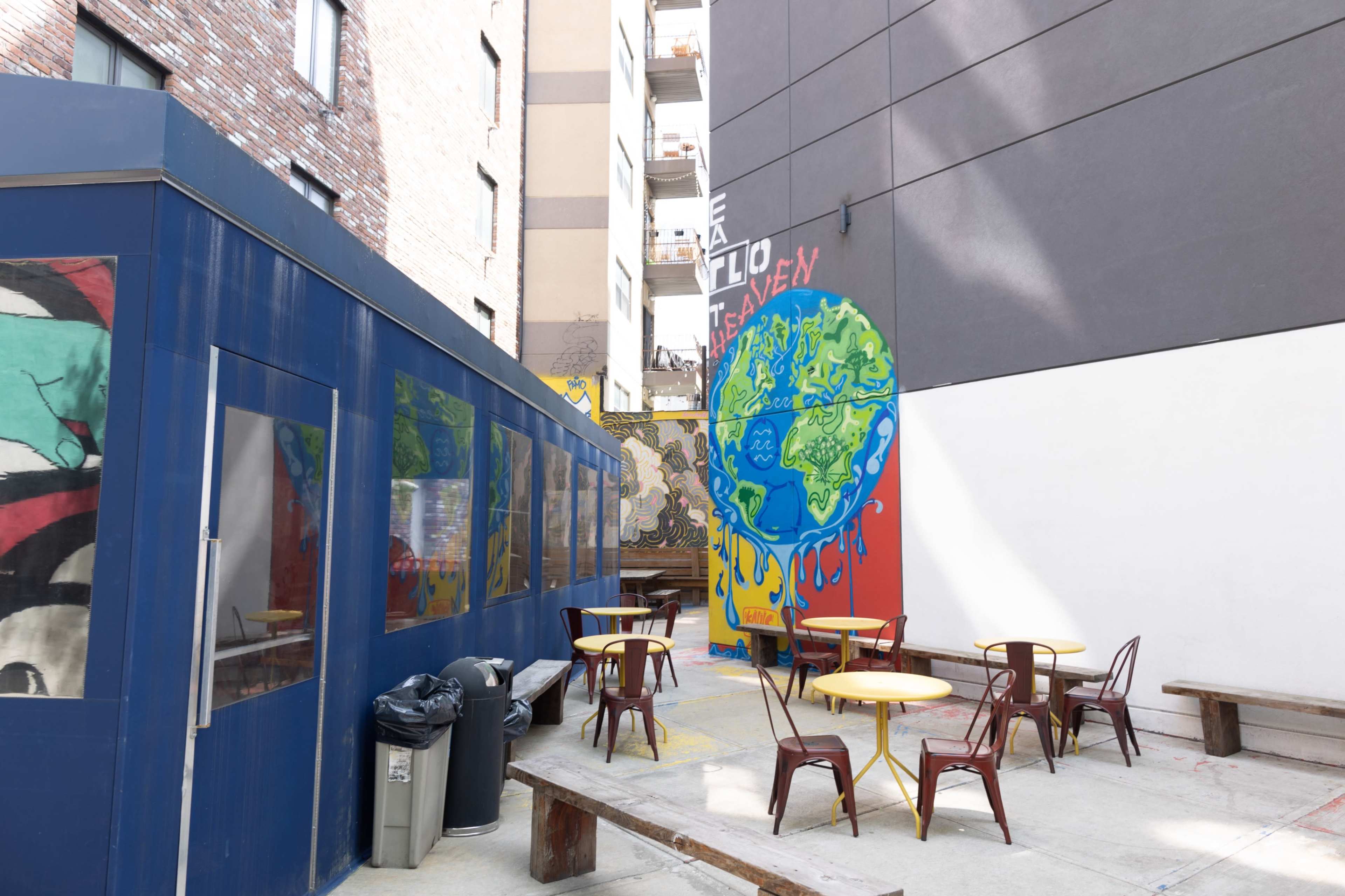 The image depicts an outdoor seating area in an urban setting, featuring colorful murals and metal tables with chairs.