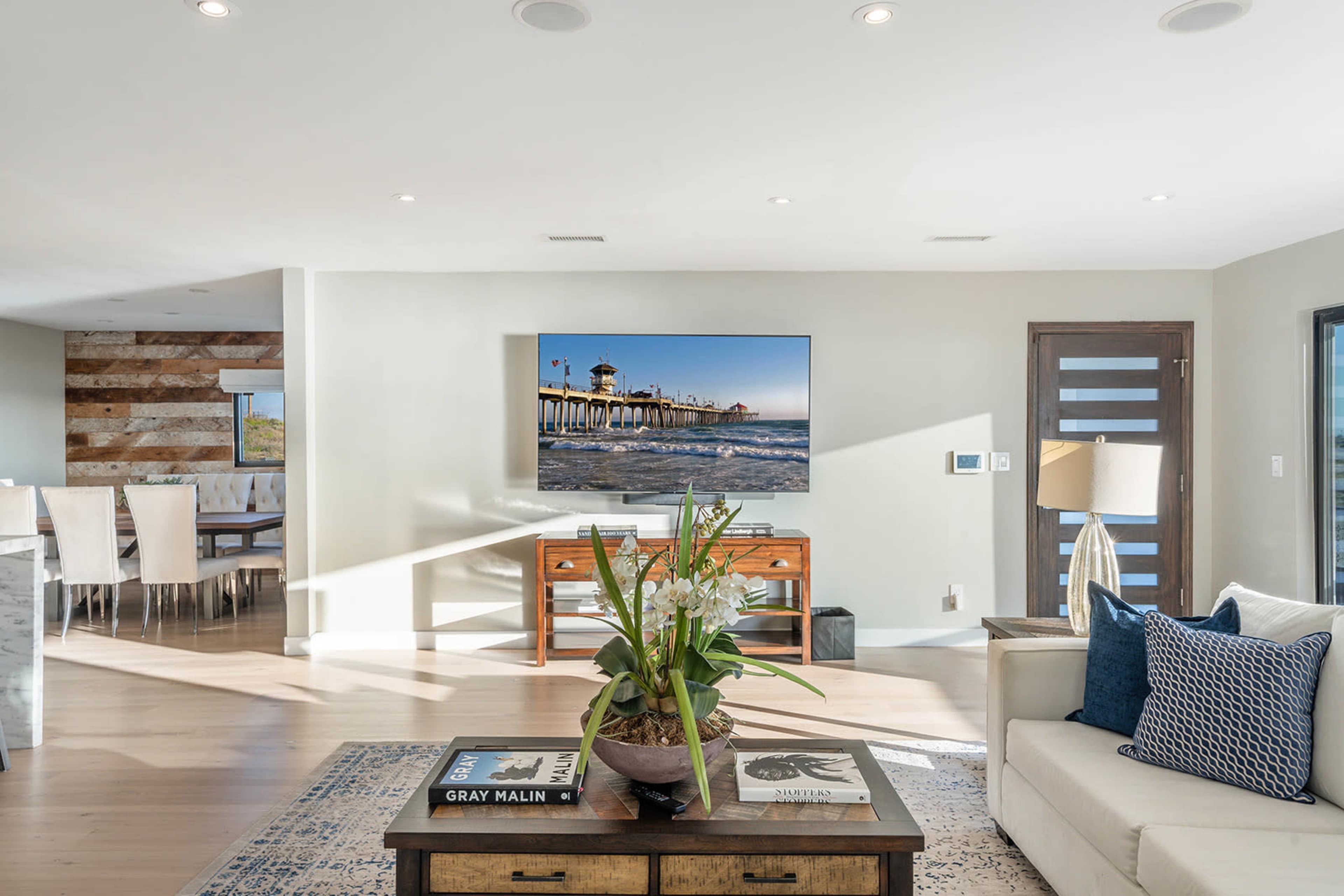 A modern living room features a light-colored sofa, a coffee table with a plant, and a large wall-mounted television displaying a coastal image.