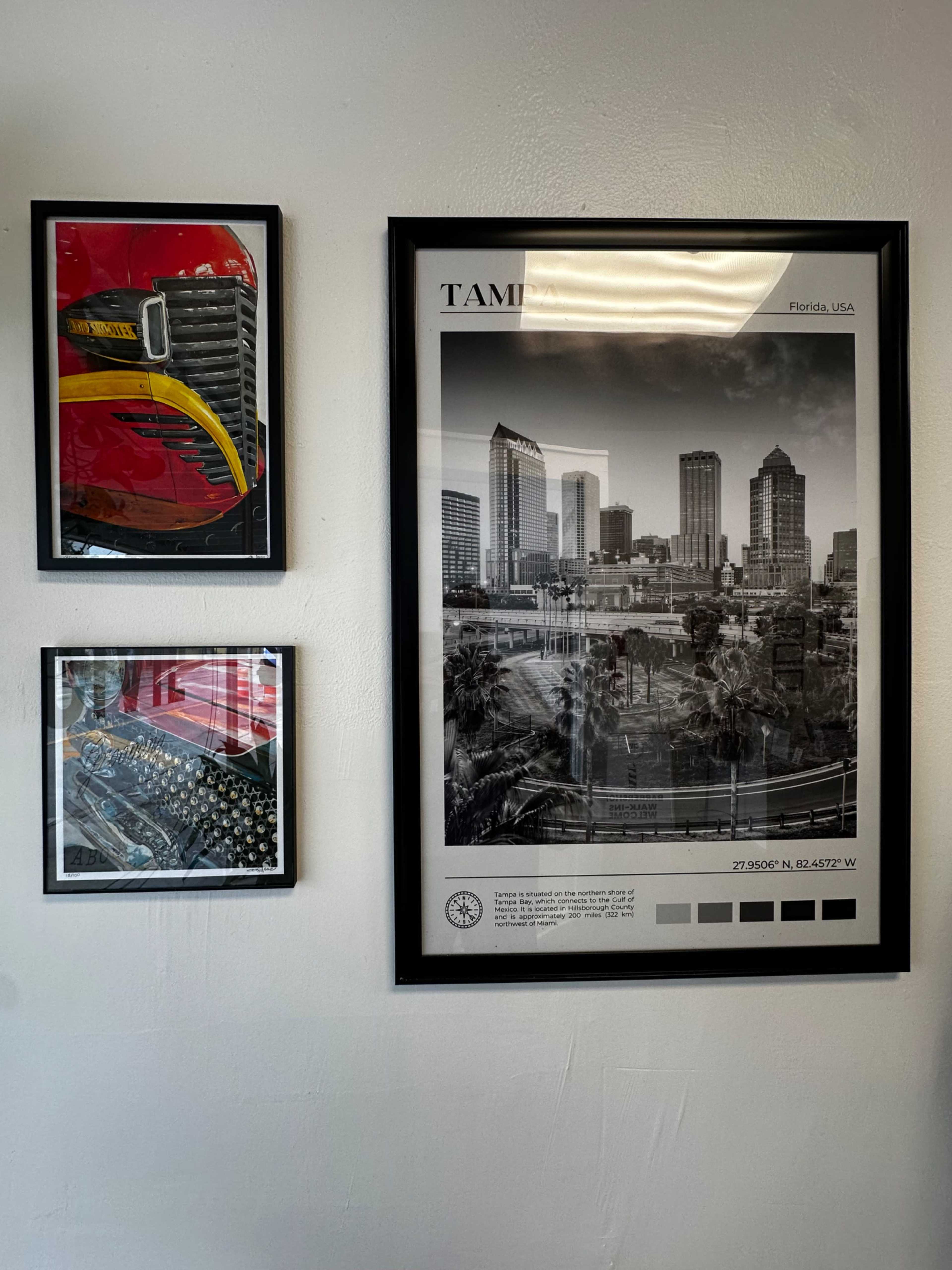 The image shows three framed artworks on a wall, featuring a large print of a city skyline, a photograph of a colorful vehicle, and a close-up of a mechanical component.