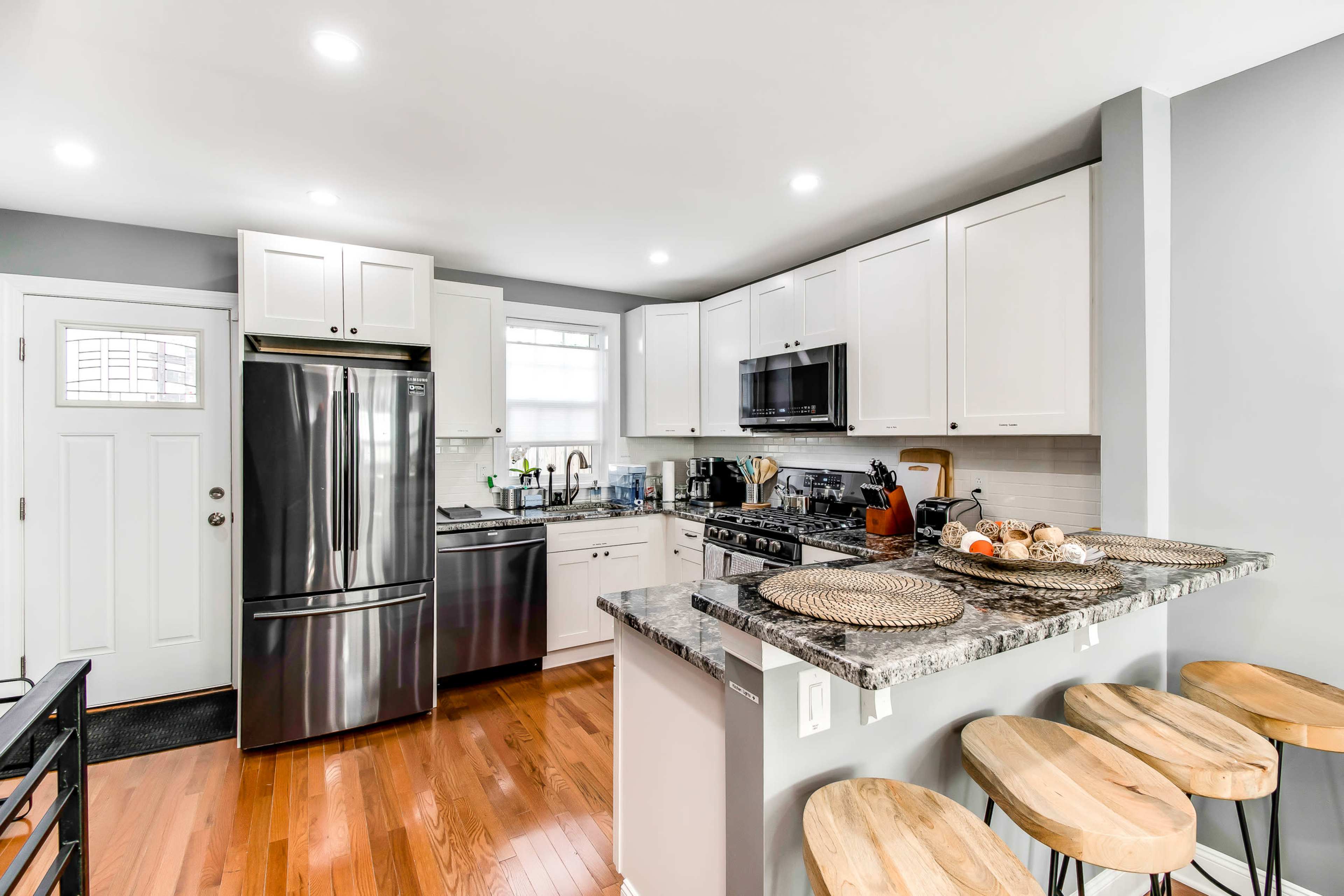 A modern kitchen features white cabinets, stainless steel appliances, and a granite countertop with wooden stools adjacent to it.
