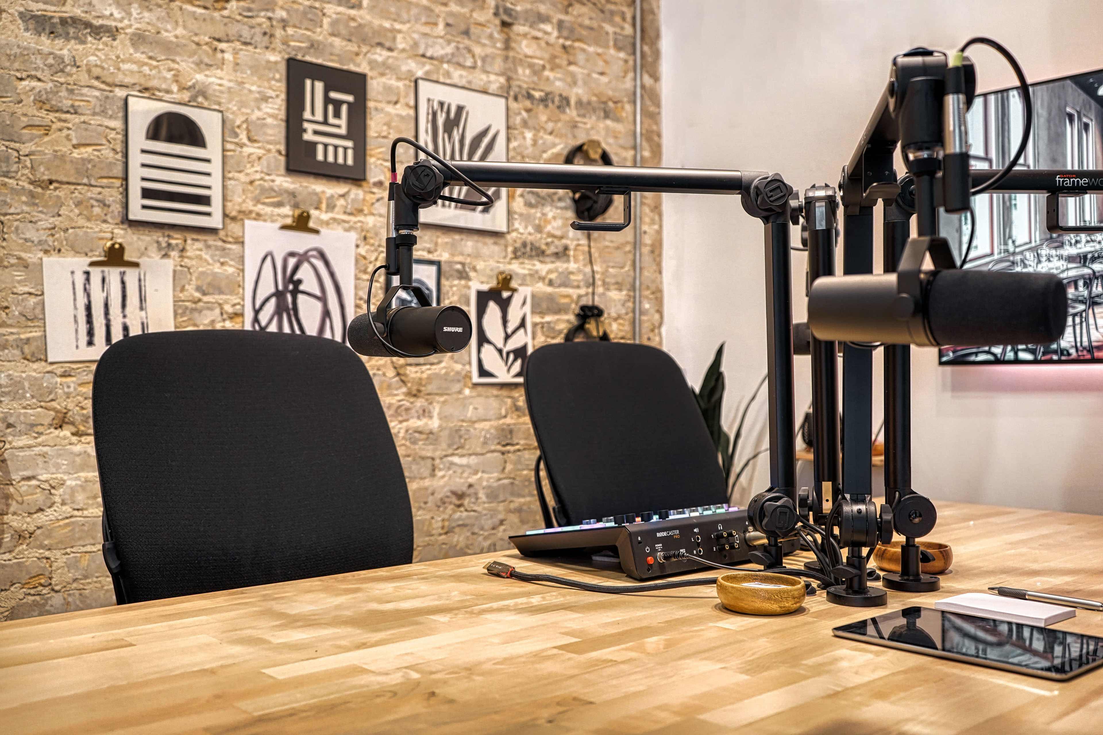 A podcast recording setup featuring two chairs, a wooden table with audio equipment, and a brick wall adorned with artwork.