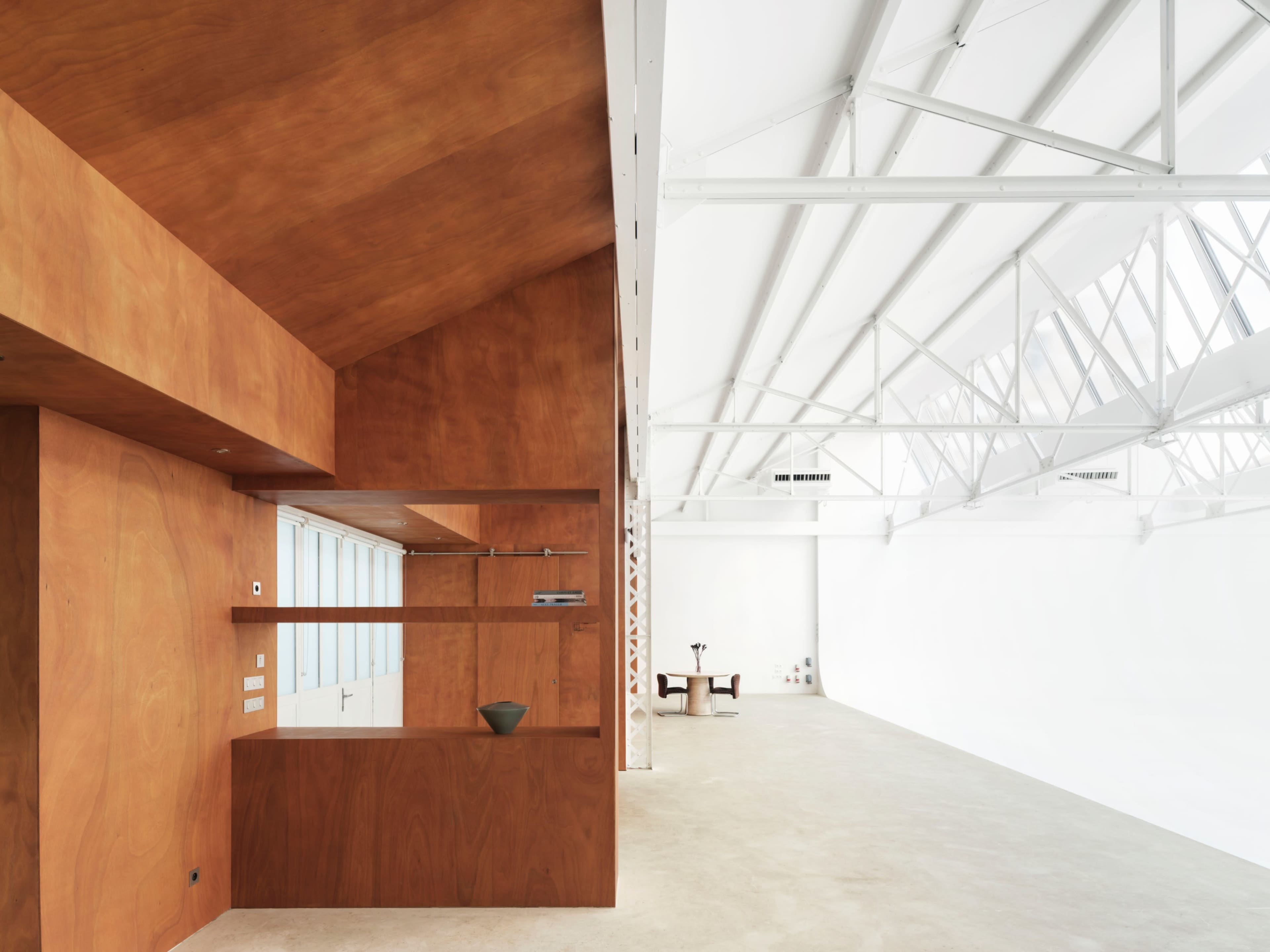 The image shows a modern interior space featuring an orange-brown wooden wall with built-in shelving on one side, contrasted by a bright, white open area with exposed beams and minimal furnishings on the other.