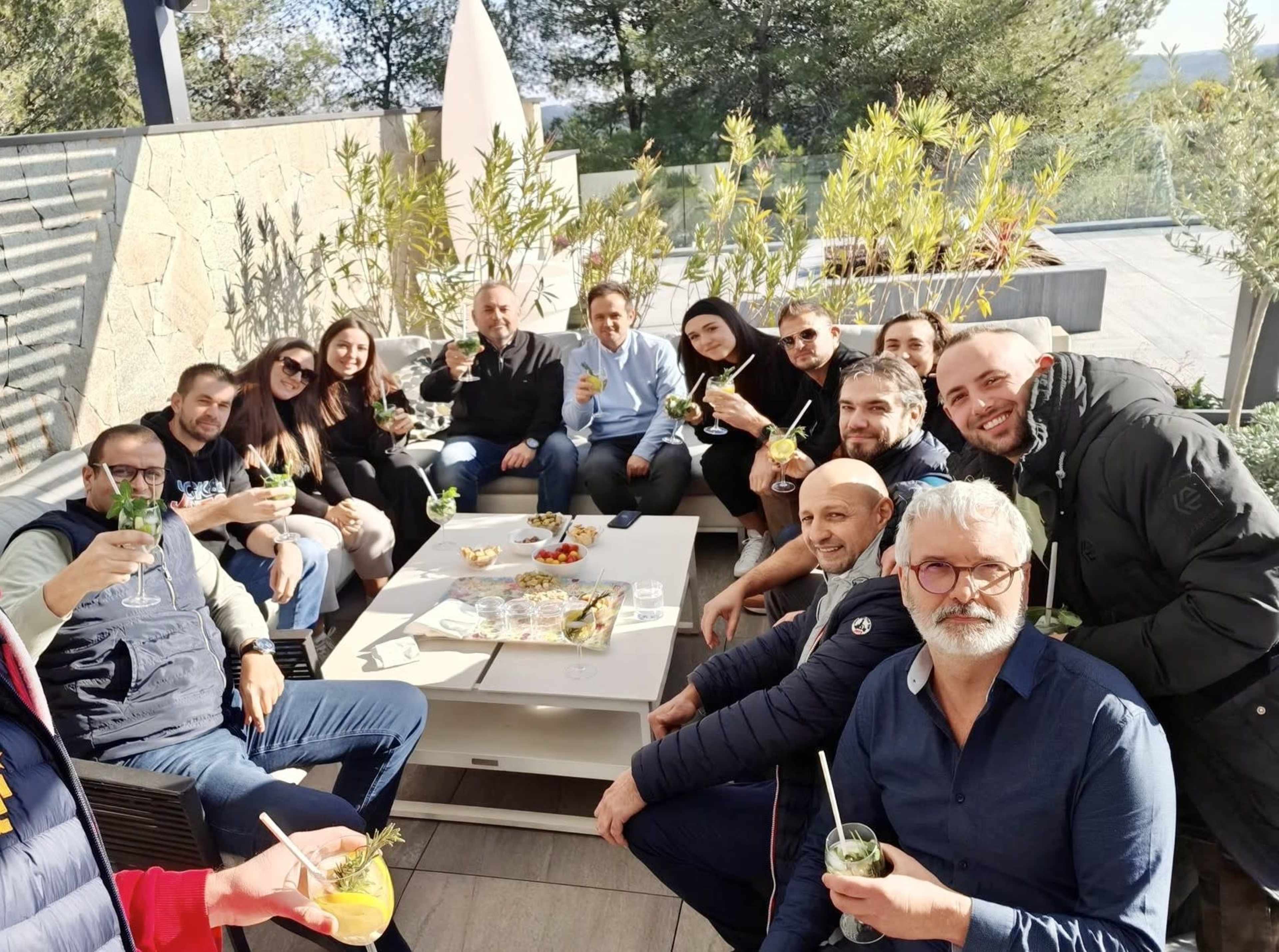 A group of people is gathered on a patio, holding drinks and smiling at the camera.