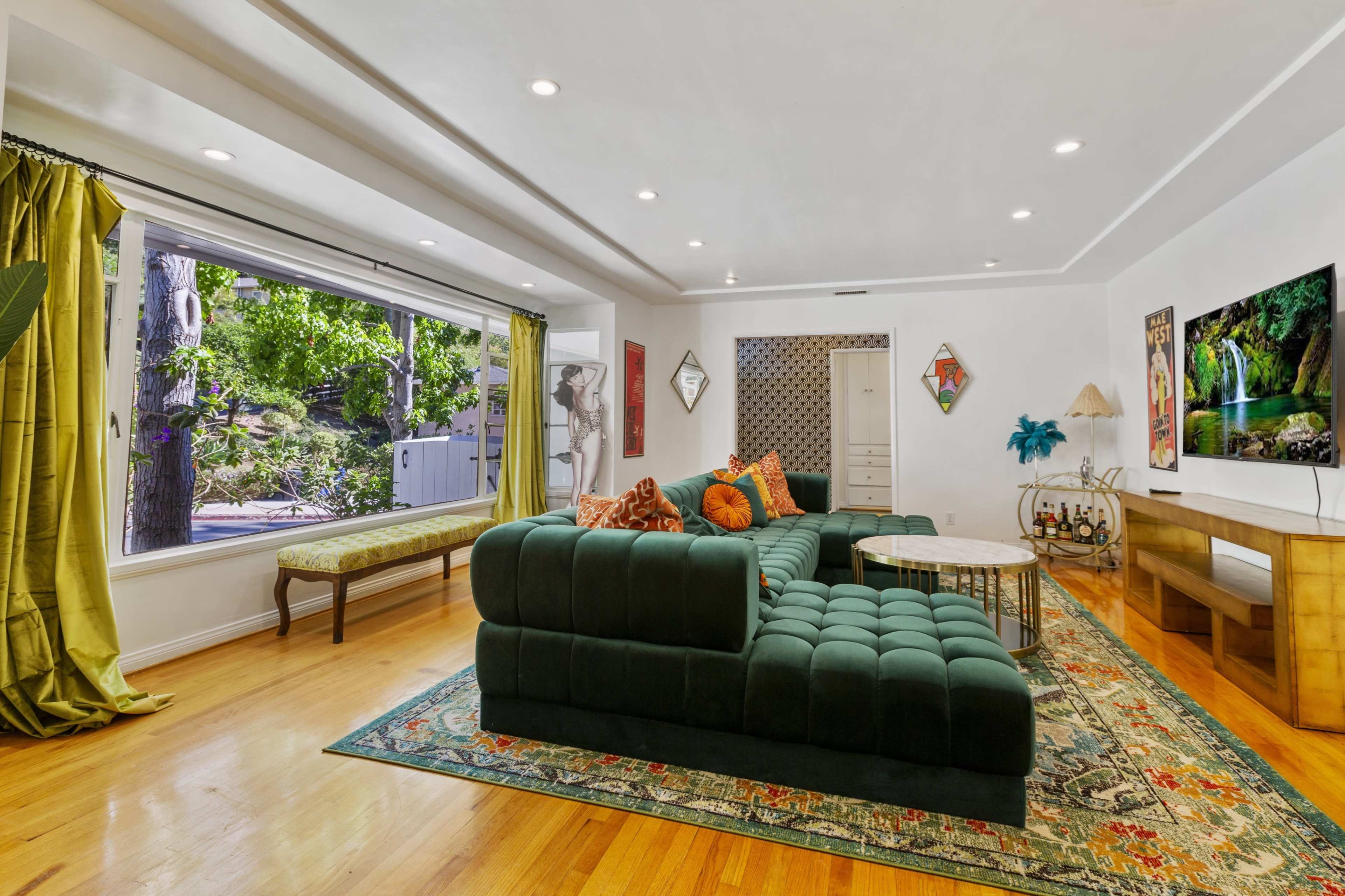 The living room features a large green sectional sofa, a coffee table, and a window that offers a view of a garden, along with art and decorative elements on the walls.