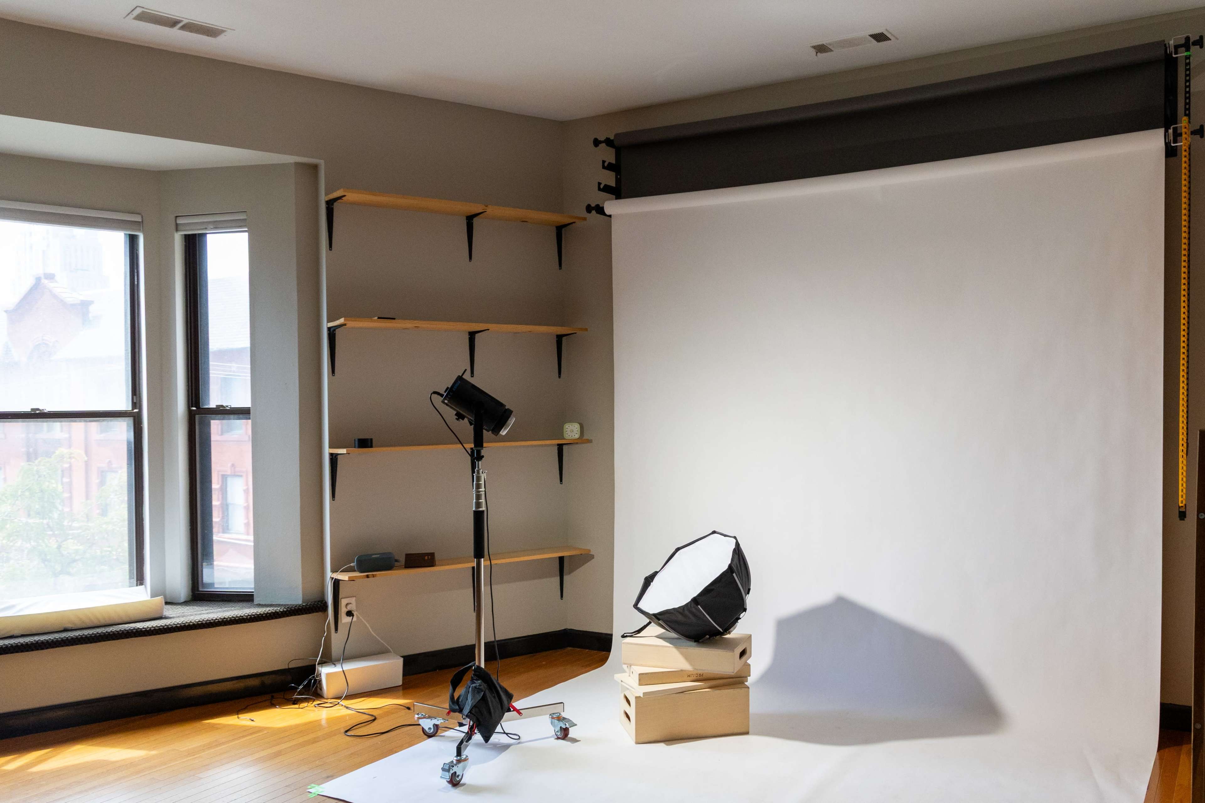 The image shows a photography studio setup with a white backdrop, a softbox light, and a tripod stand surrounded by boxes on a wooden floor.