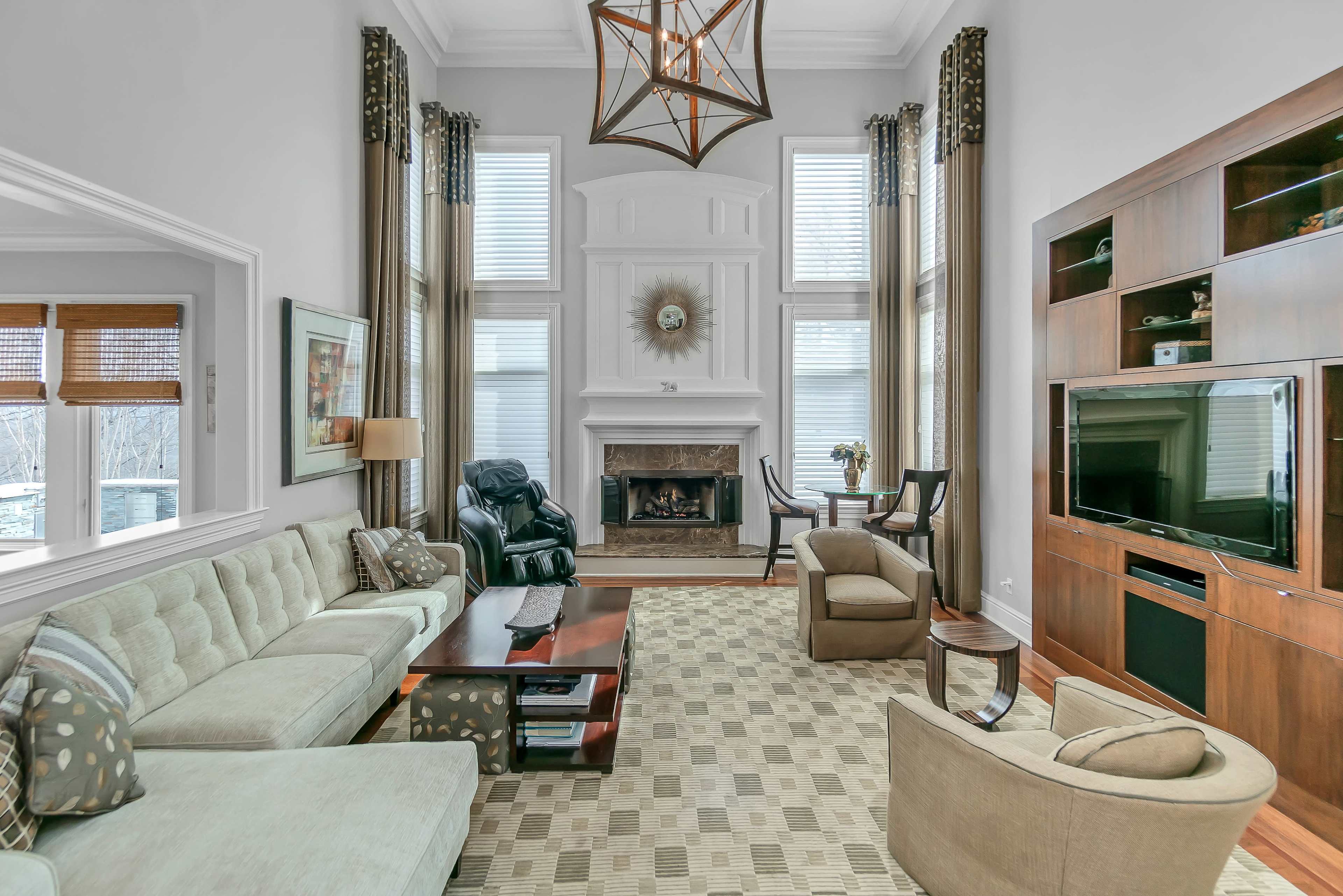 The living room features a sofa, recliner, and armchair arranged around a coffee table in front of a fireplace, with large windows allowing natural light and a television mounted on a wooden panel.