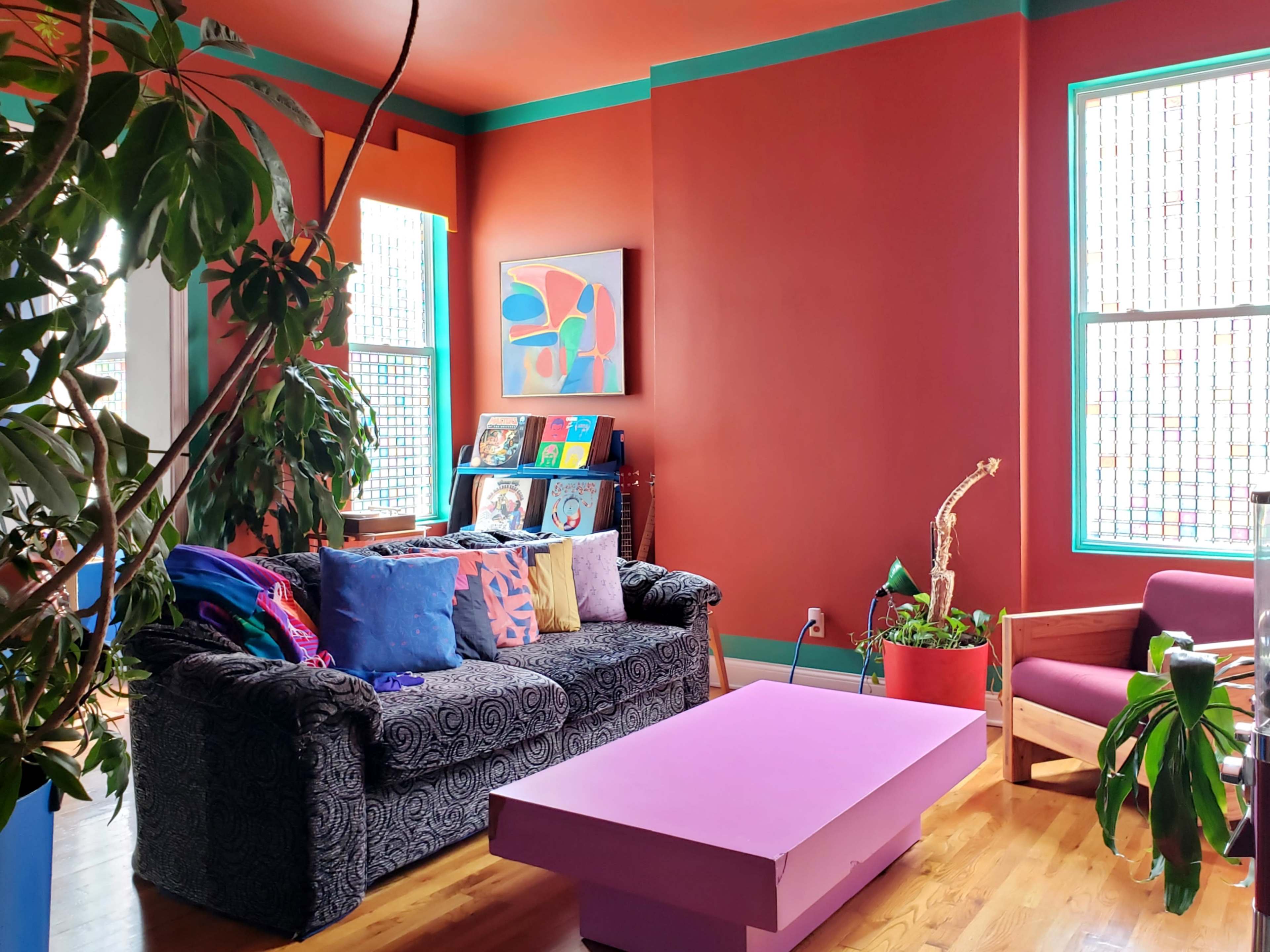 The scene depicts a bright, modern living room with a dark patterned sofa, colorful pillows, a purple coffee table, and large windows featuring stained glass.