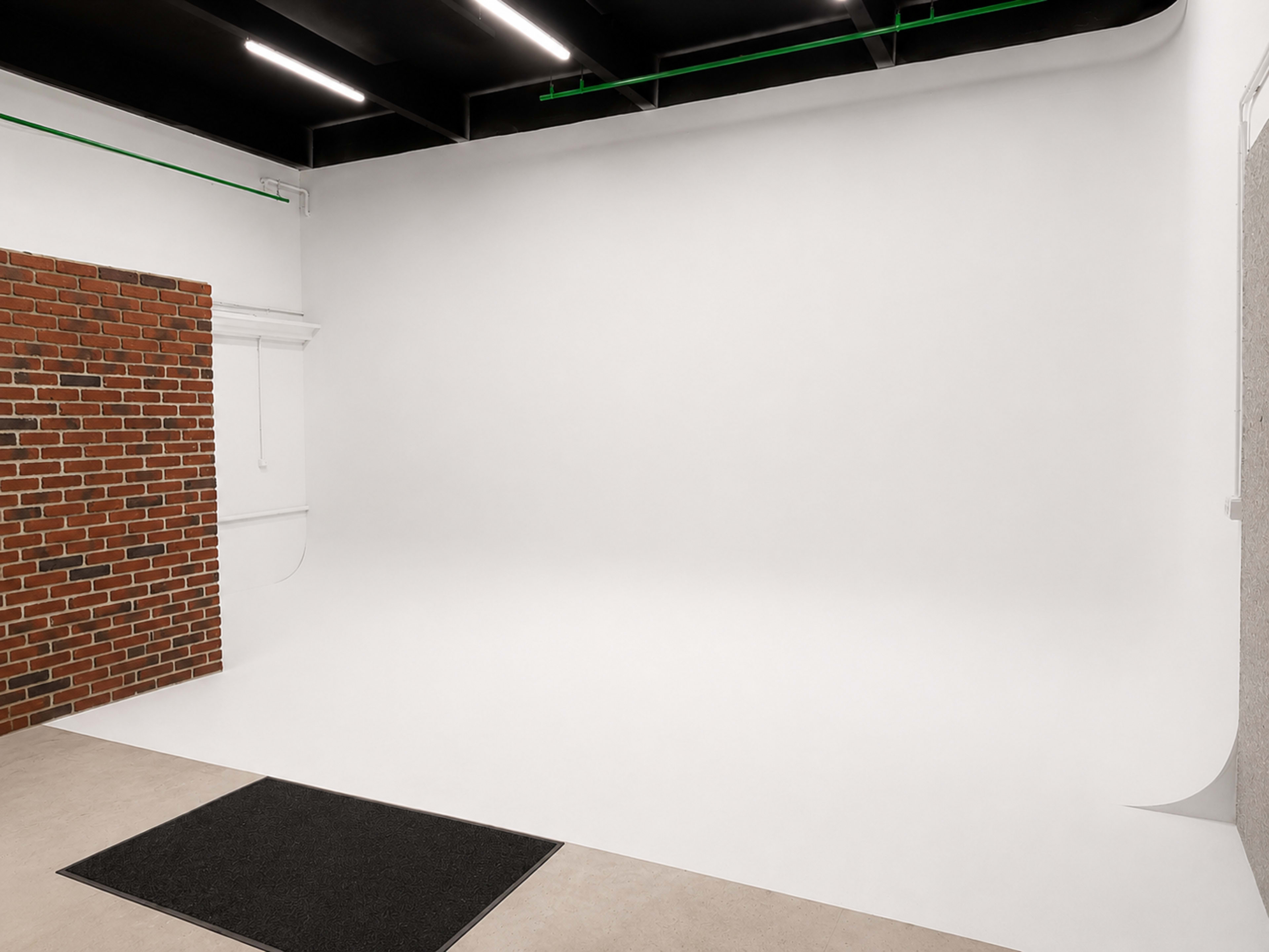 The image shows a photography studio featuring a smooth white backdrop, a section of exposed red brick wall, and a light-colored floor mat near the entrance.