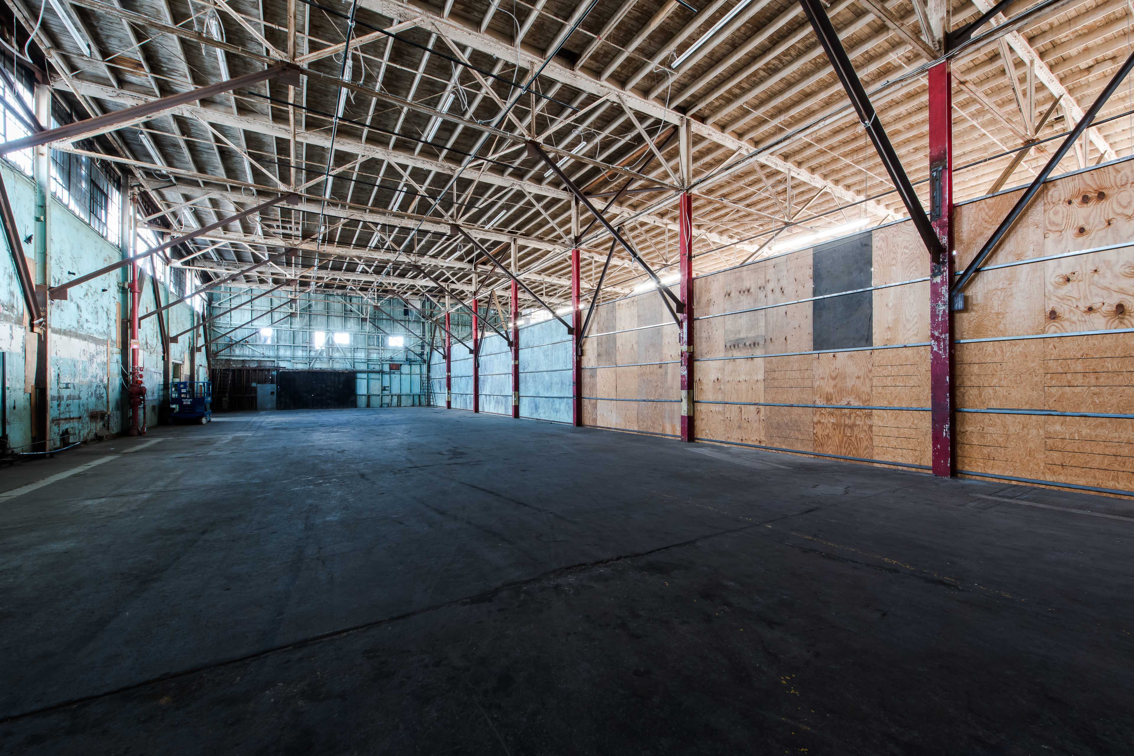 The image shows a spacious, empty warehouse with wooden beams, concrete flooring, and partially boarded walls.