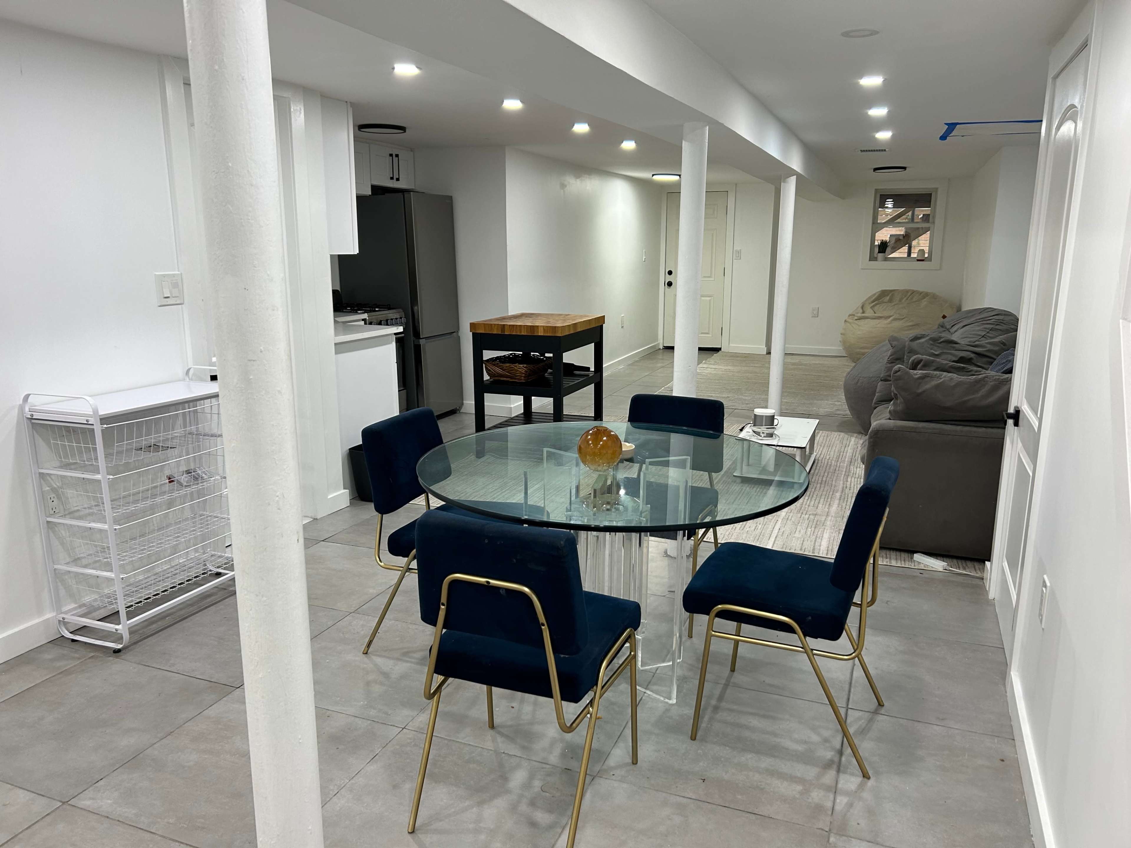 The image shows a modern basement interior with a glass dining table surrounded by four blue chairs, a wooden table, a kitchen area, and a large bean bag in one corner.