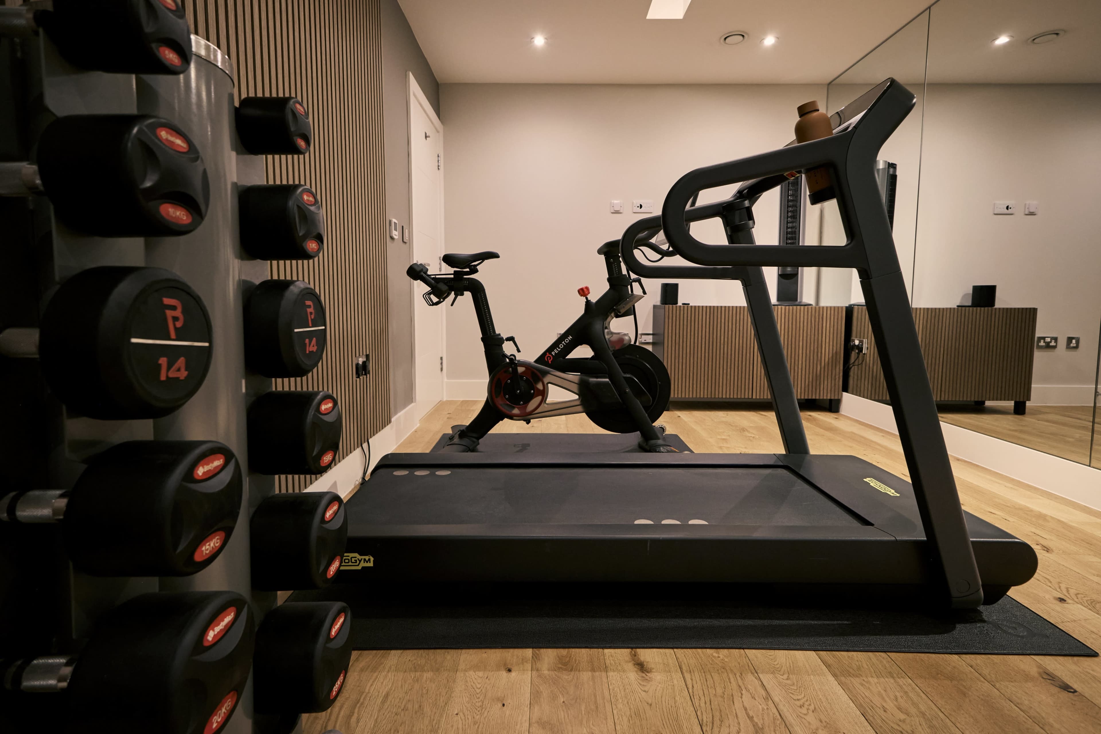 The image shows a modern fitness room featuring a treadmill, a stationary bike, and a row of dumbbells.