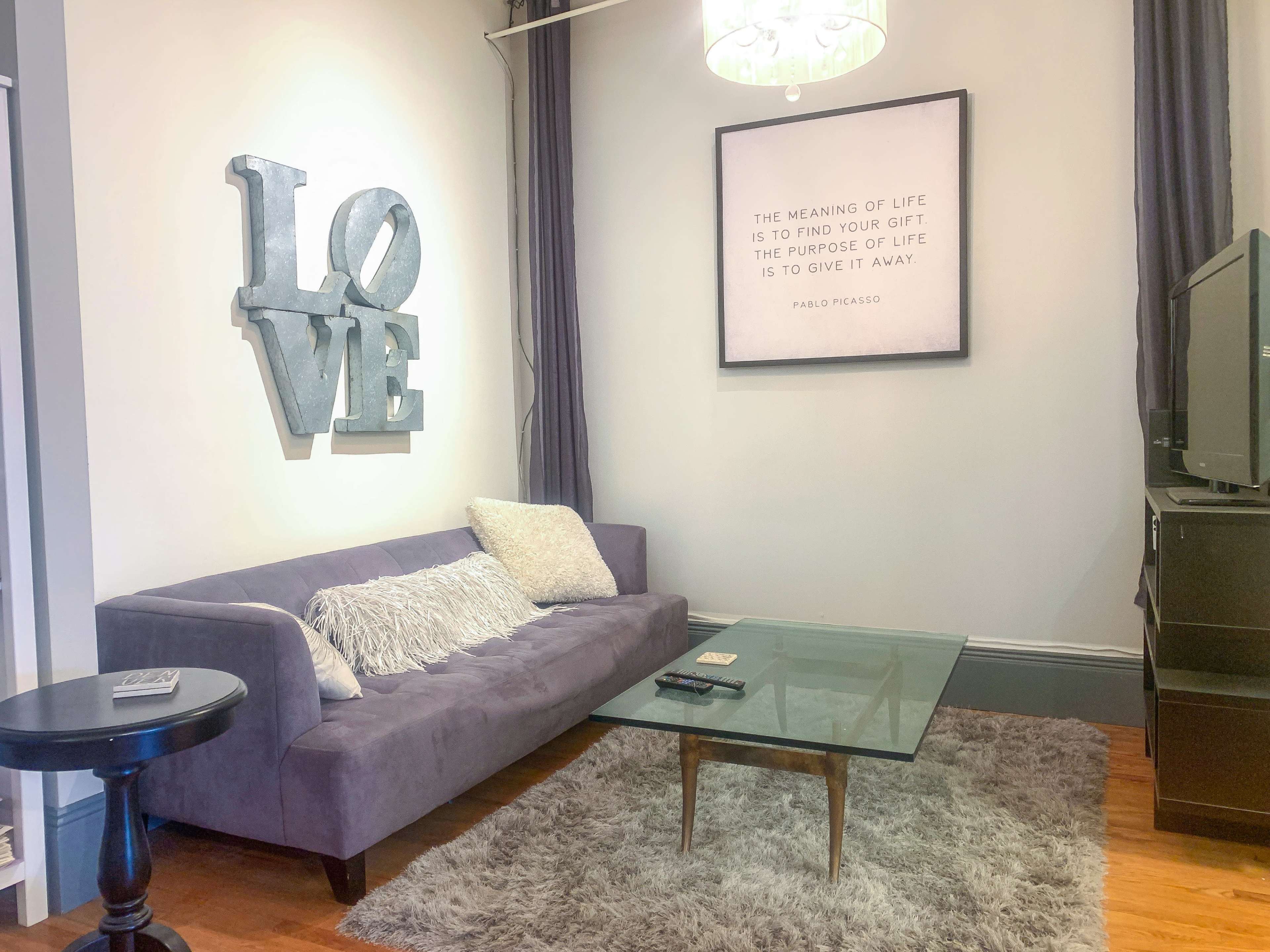 A purple couch with a decorative throw pillow sits in front of a glass coffee table on a plush rug, next to a wall featuring the word "LOVE" in large letters and a framed quote by Pablo Picasso.