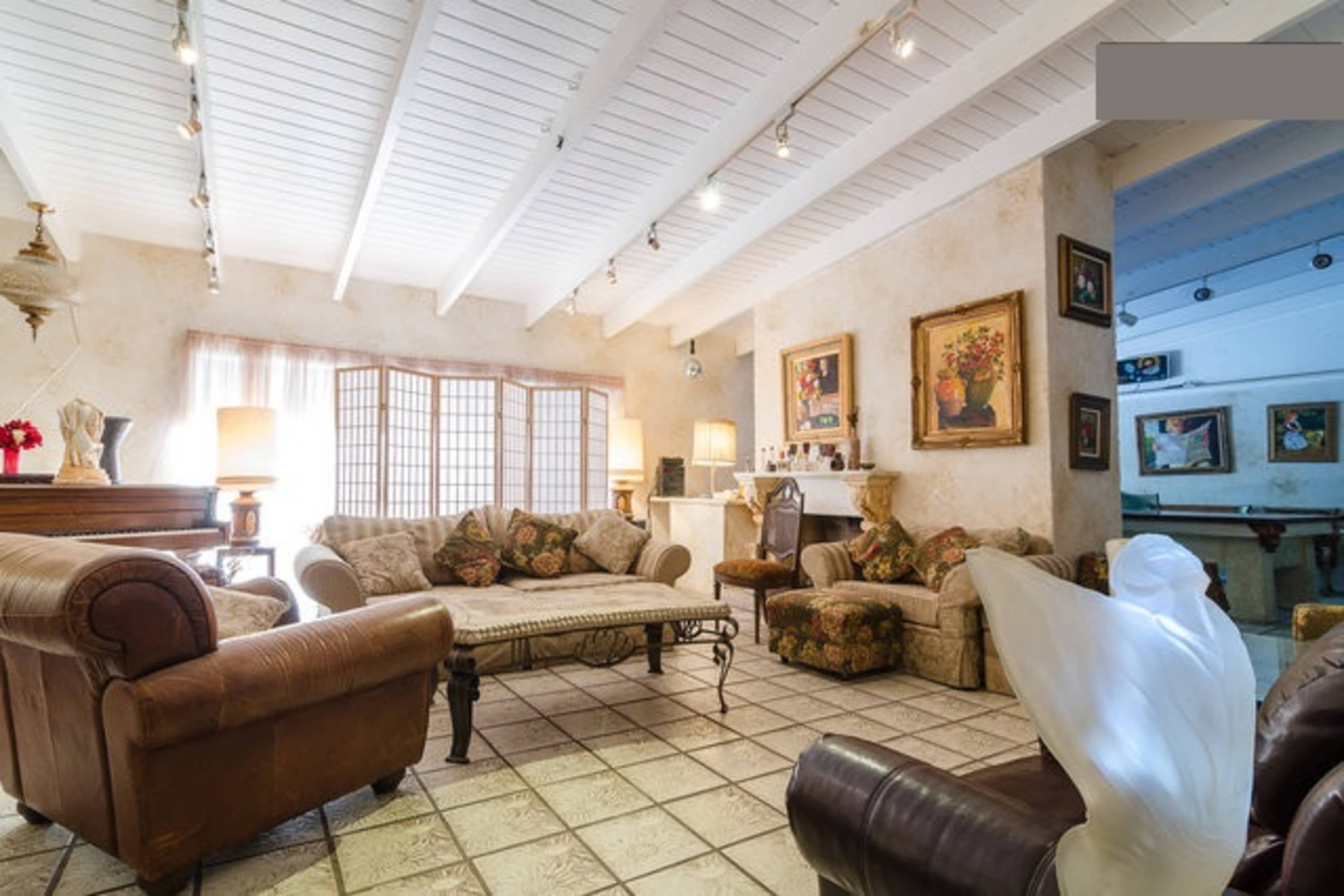 A spacious living room with a patterned sofa, several artworks on the walls, and a large window allowing natural light to enter.