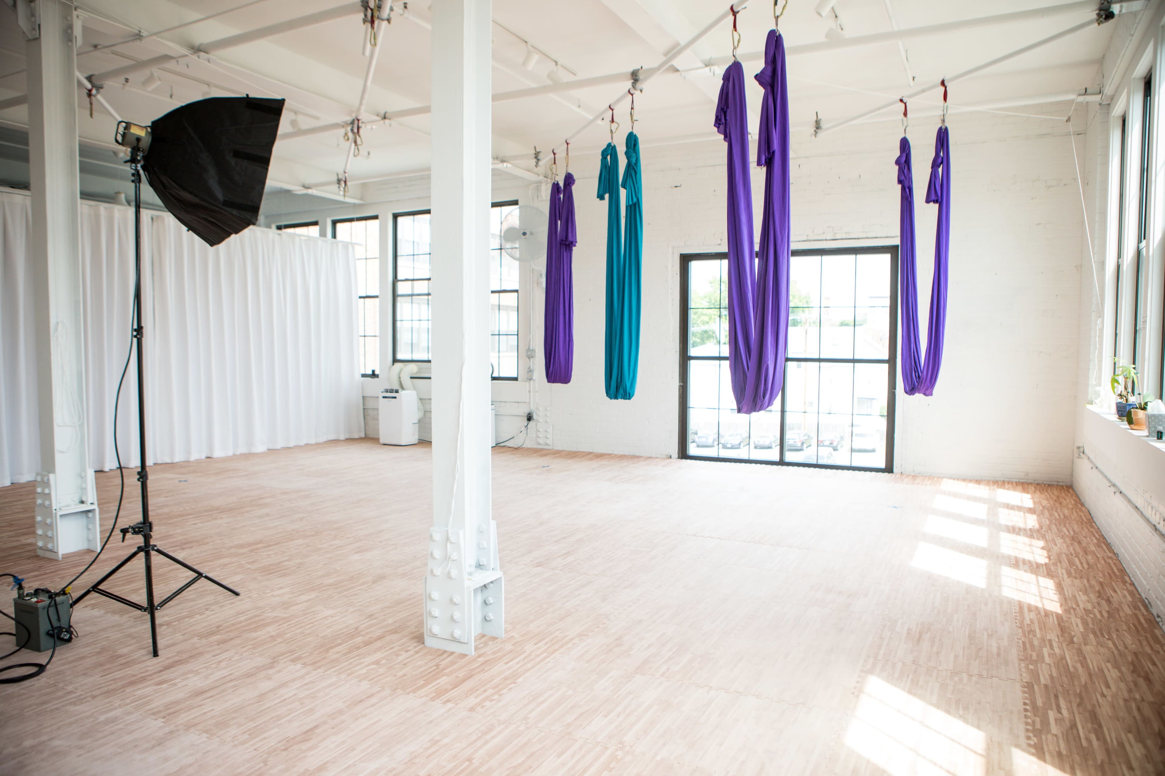 The image shows a spacious, well-lit studio with large windows, featuring aerial silk hangings in purple and blue colors and a photography light setup.