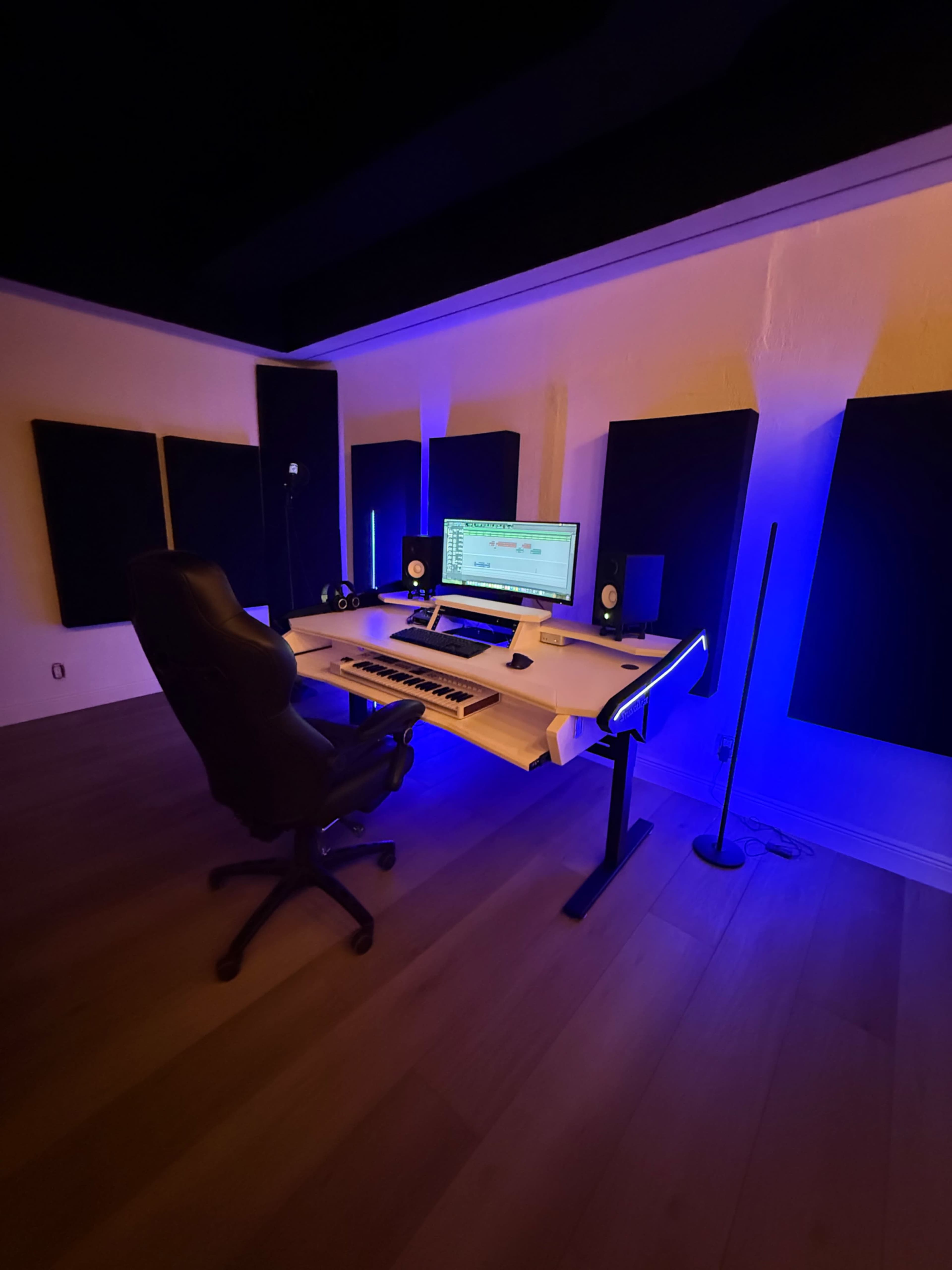 The image shows a music production studio featuring a desk with a computer, speakers, and acoustic panels, illuminated by blue light.