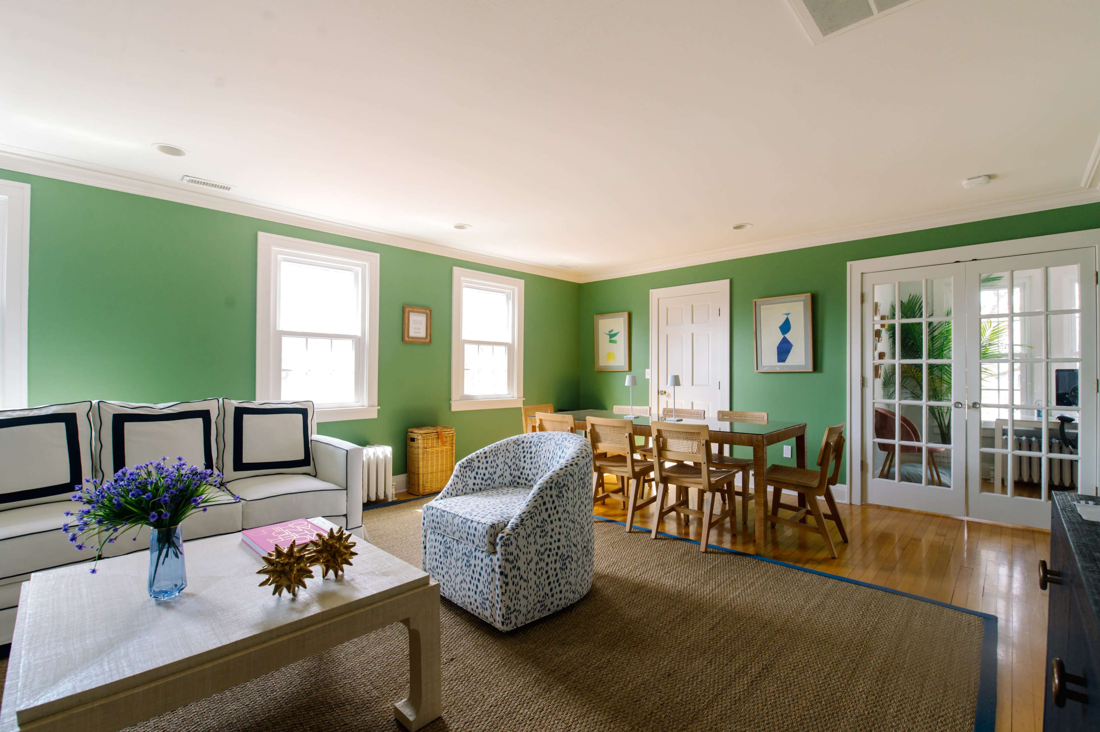 The image shows a bright living room with green walls, a white sofa, a blue and white patterned chair, a wooden dining table, and a natural fiber rug.