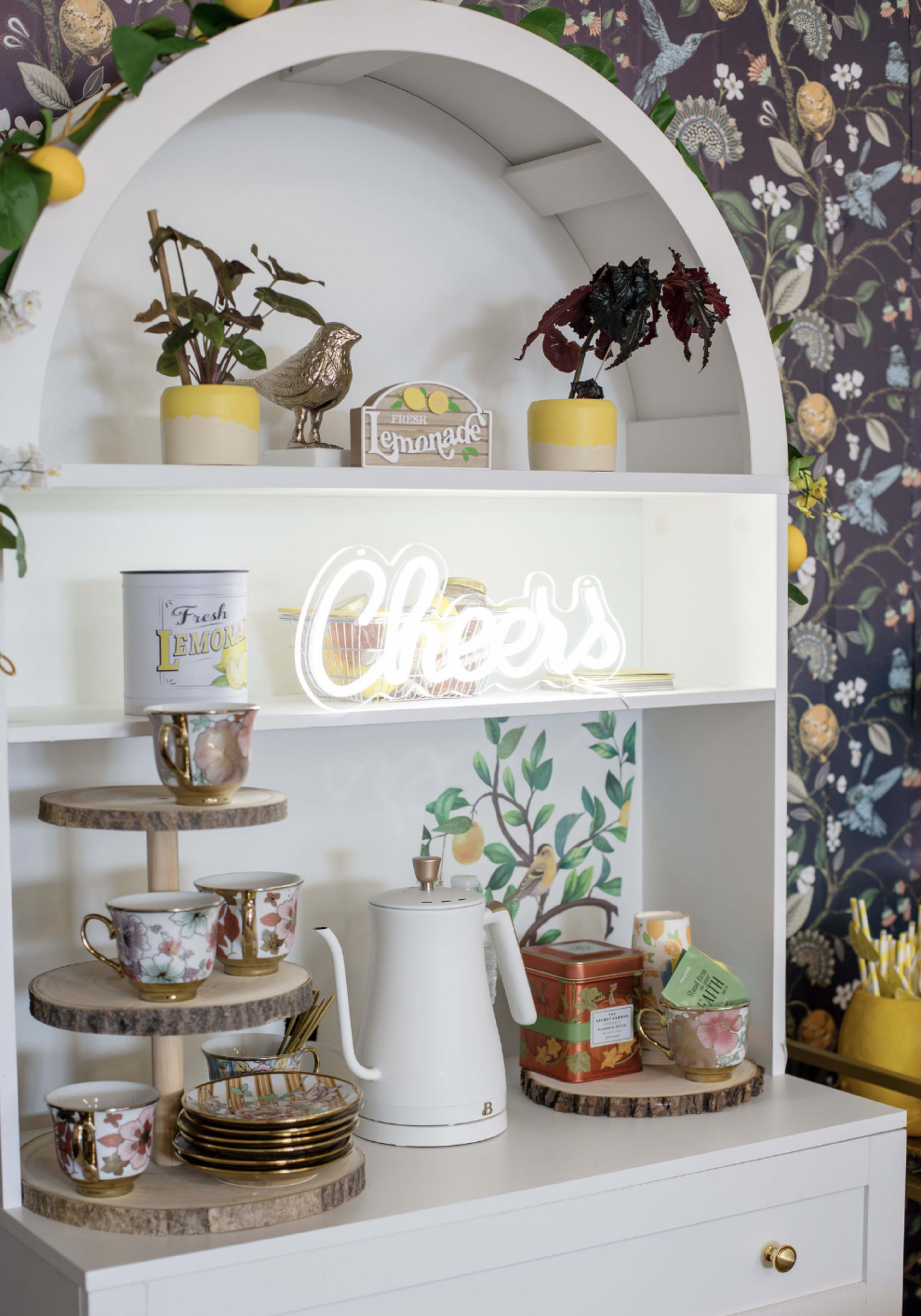 The image shows a white shelf with decorative items, including a neon sign that says "Cheers," various cups, a teapot, and potted plants.