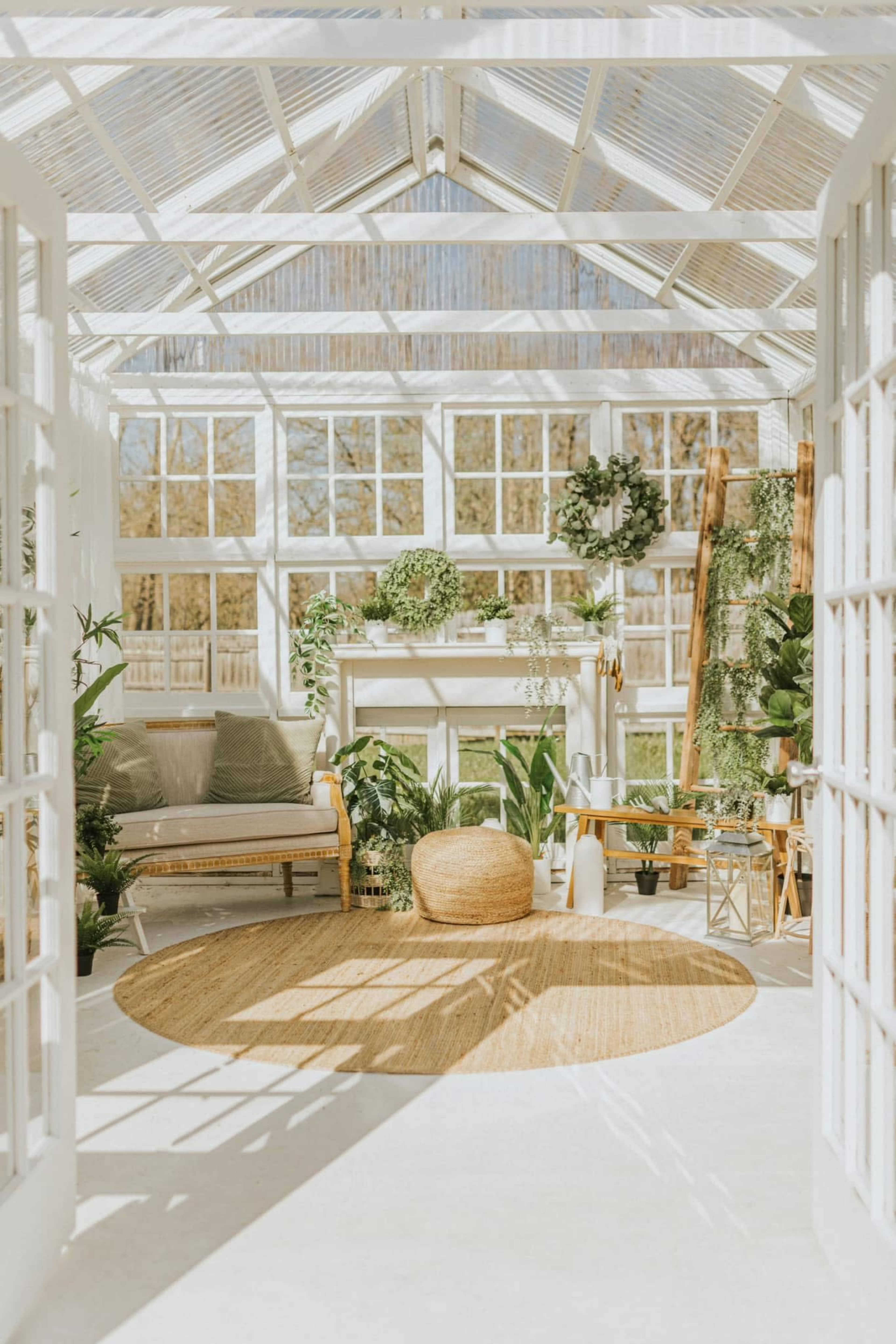 The image shows a bright, airy greenhouse filled with plants, a cozy seating area, and large windows allowing natural light to illuminate the space.
