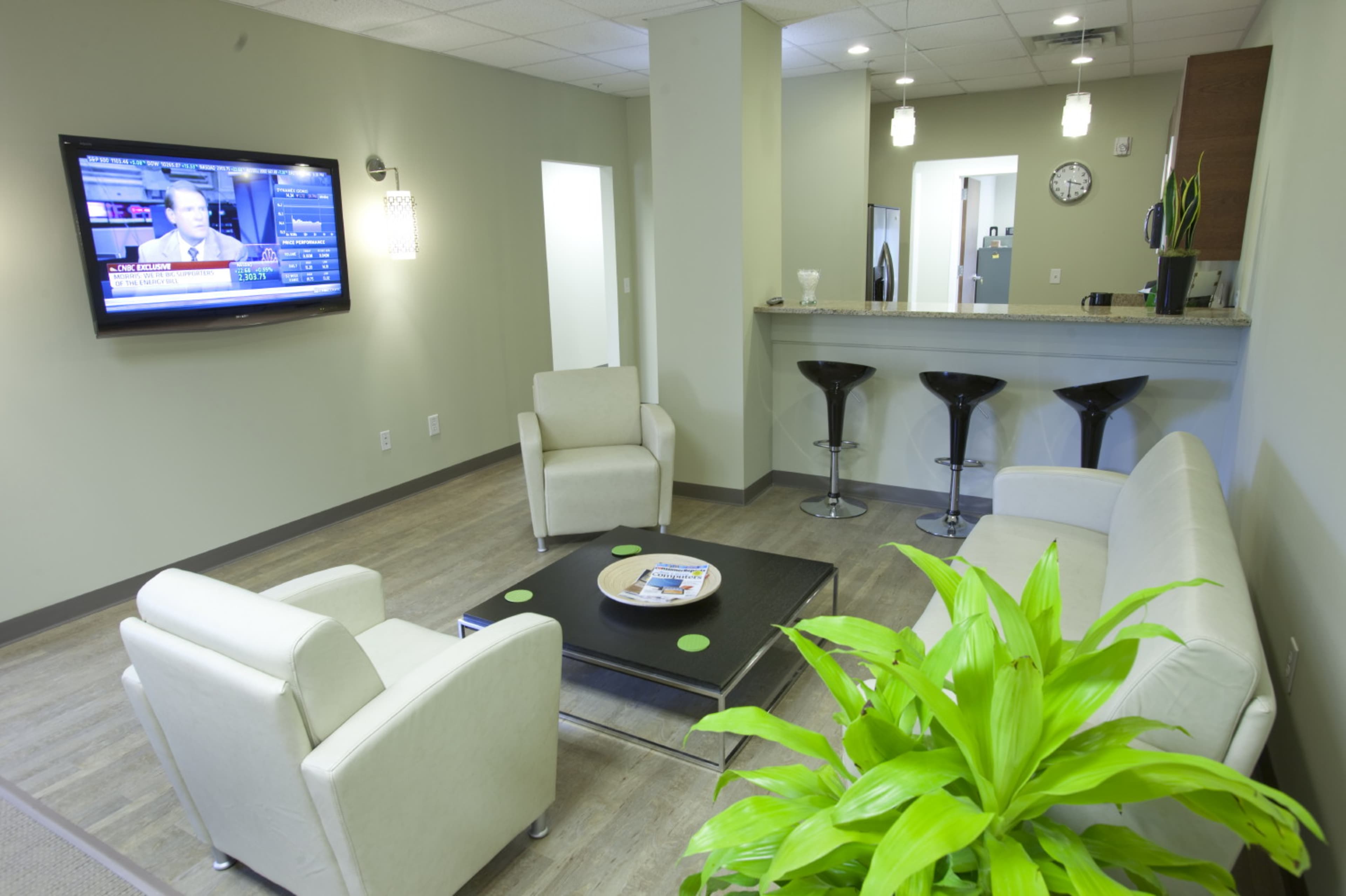 A modern waiting area with two light-colored sofas, a coffee table, a television on the wall, and a bar counter with black stools in the background.