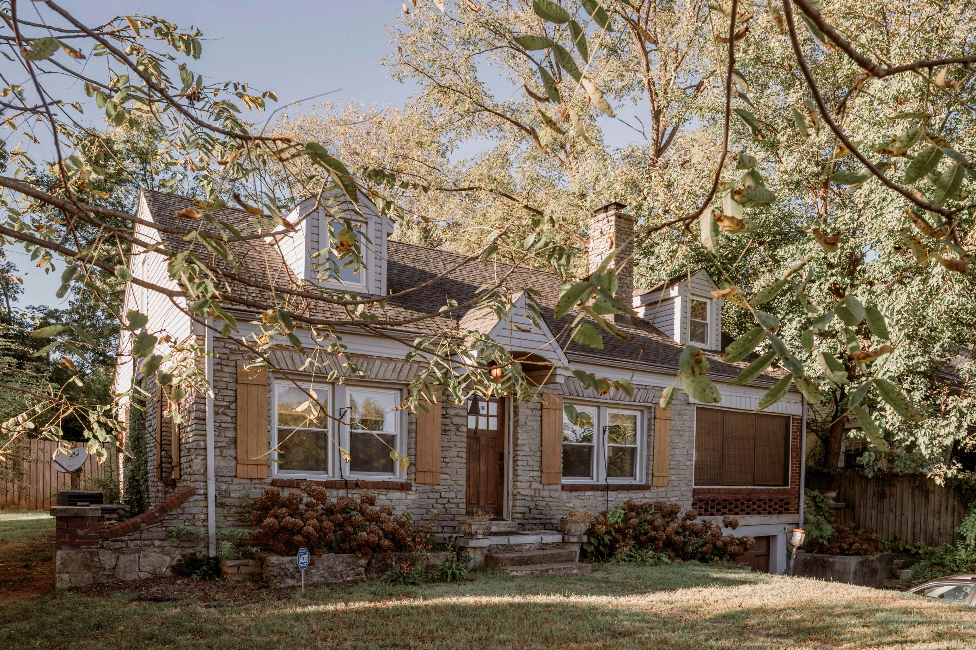 The image shows a two-story house with a stone exterior, located in a garden with trees and shrubs.