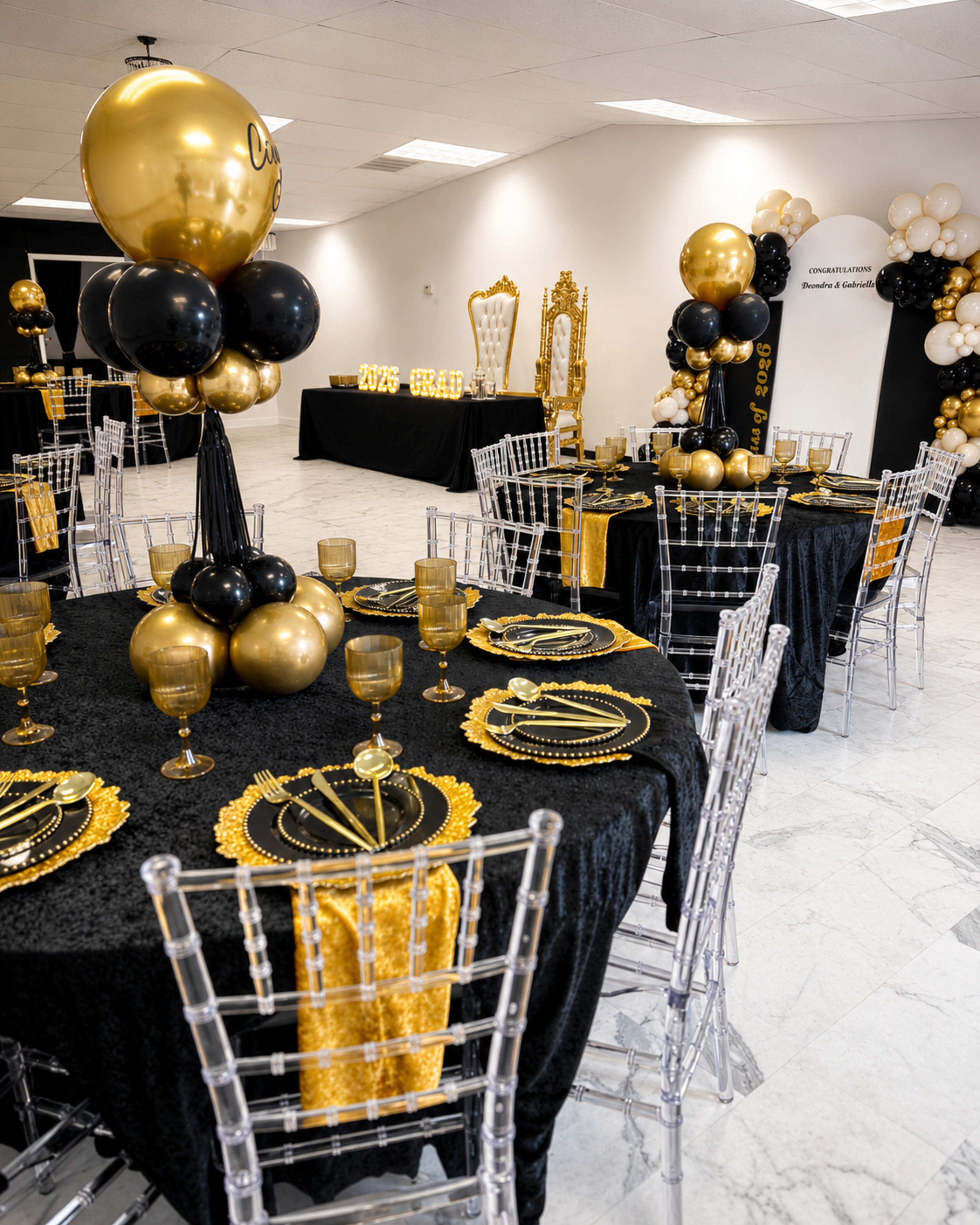 The image shows a decorated event space with black and gold table settings, featuring elegant tables with sheer chairs, gold balloons, and decorative centerpieces.
