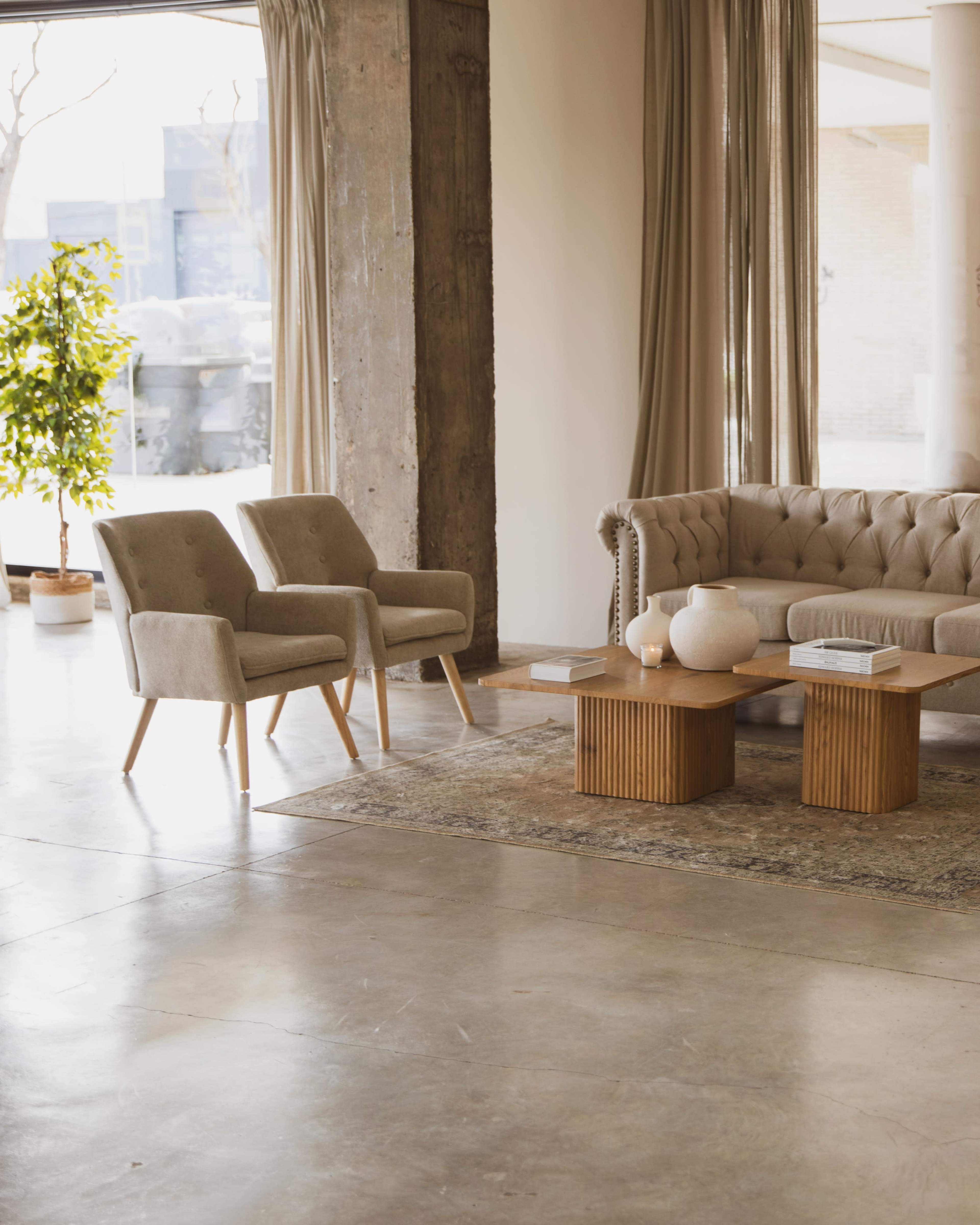 A modern living room features two gray armchairs, a tufted sofa, and wooden coffee tables on a patterned rug, with large windows allowing natural light to enter the space.
