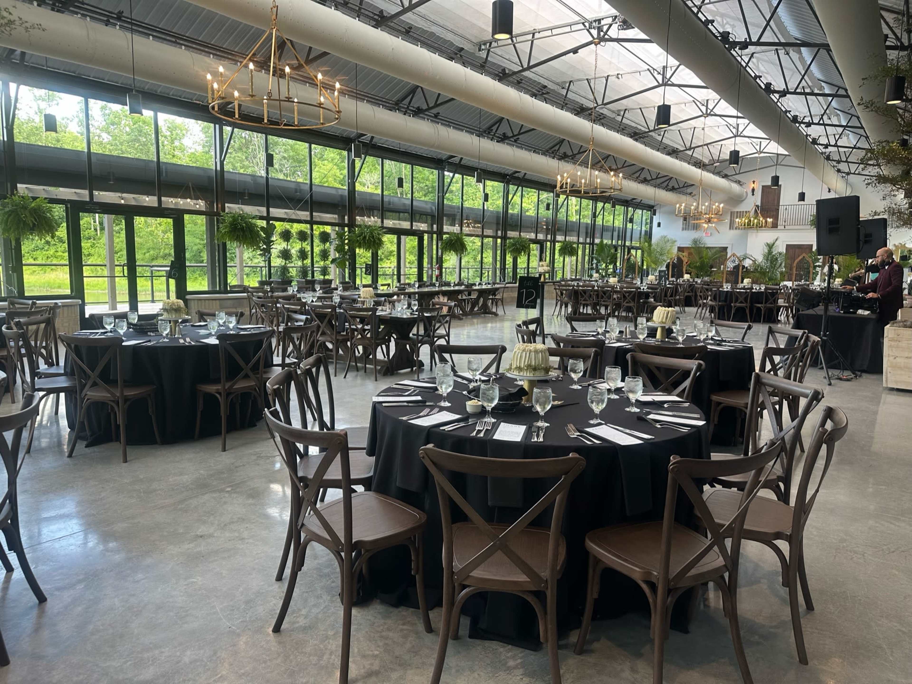 The image shows a spacious dining area with round black tables set for a formal event, surrounded by large windows that offer a view of green trees outside.