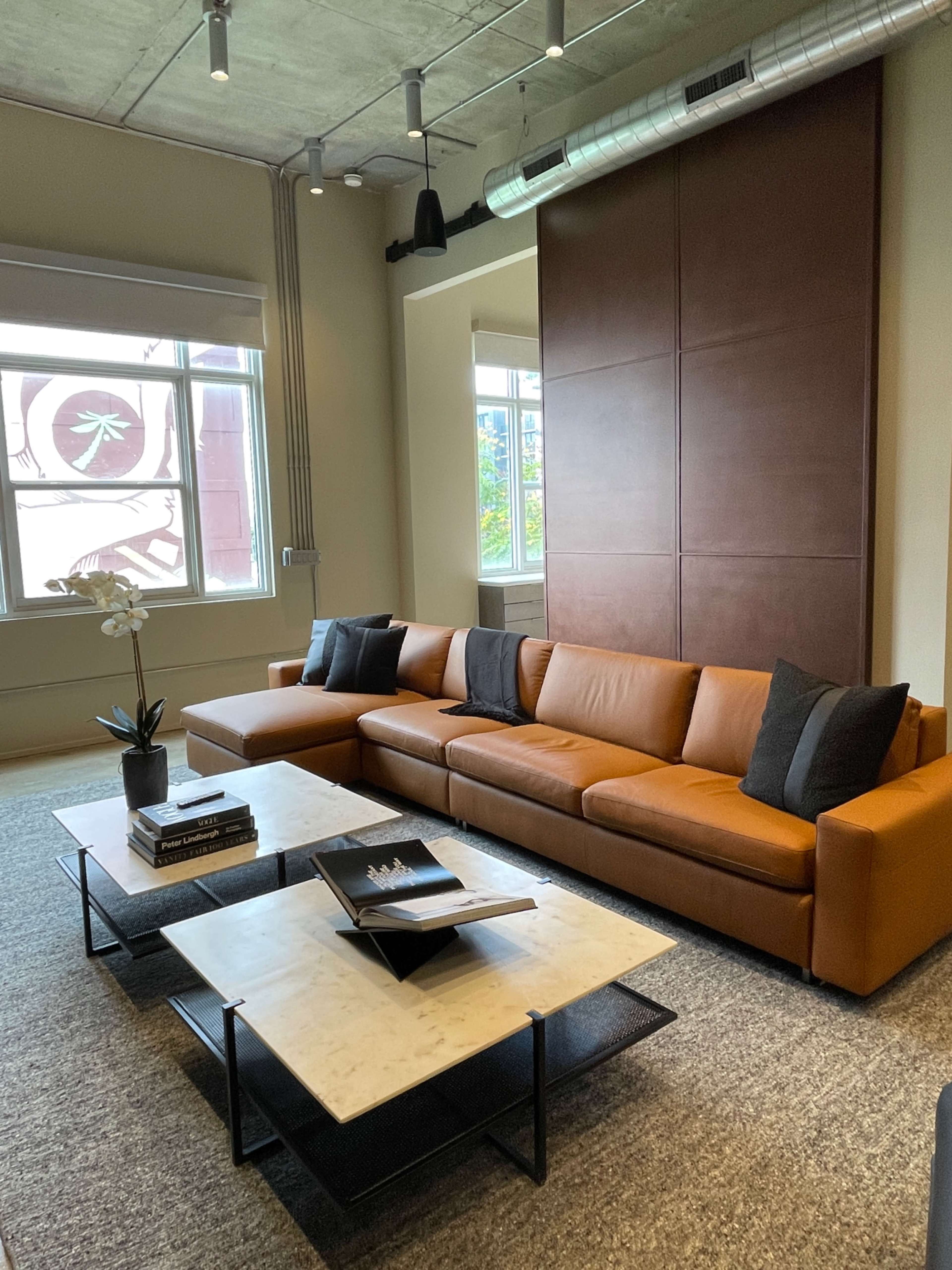 A modern living room featuring a large brown leather sofa with black cushions, a marble-topped coffee table, and large windows allowing natural light to enter.