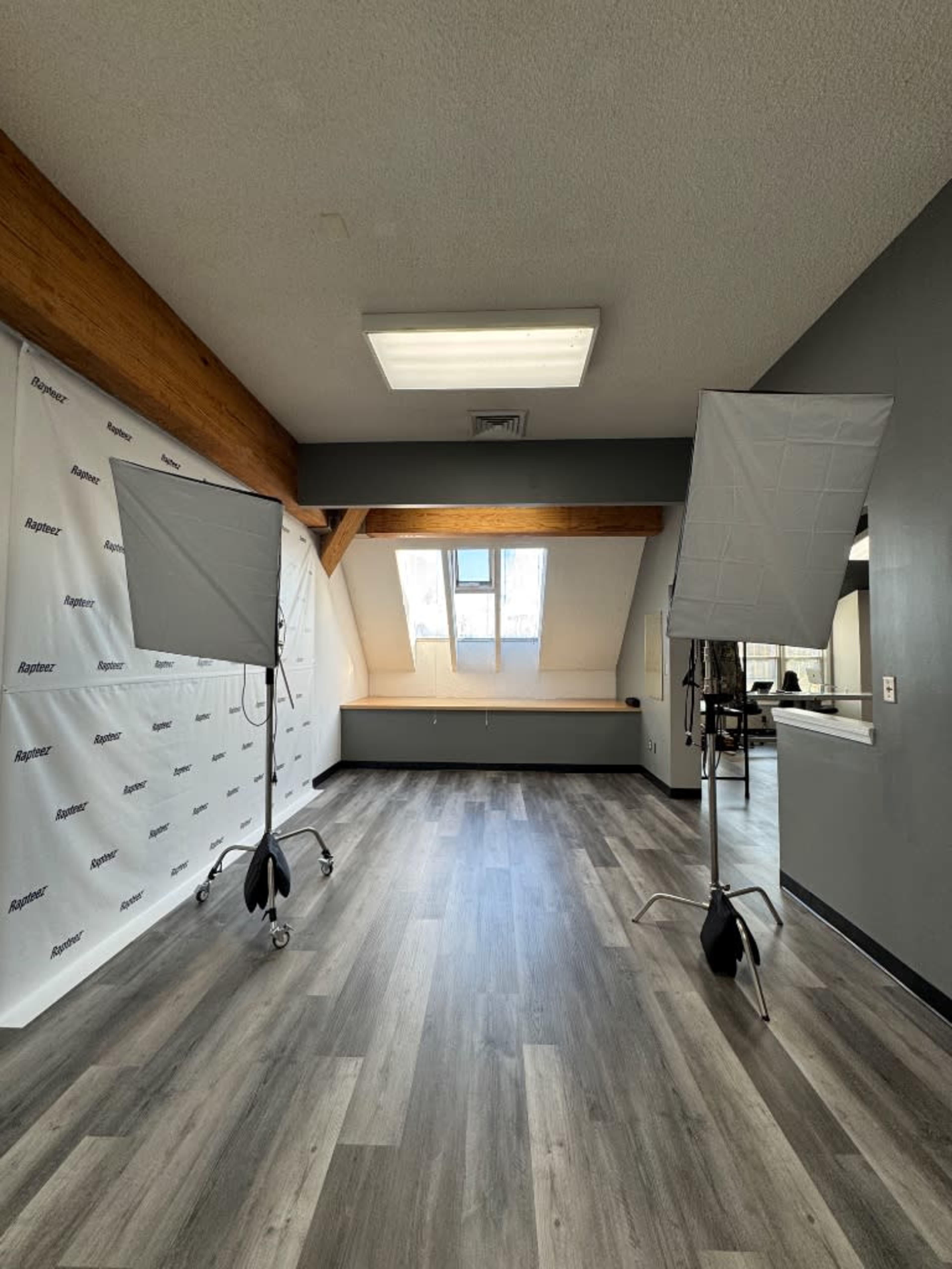 The image shows a spacious area with wooden beams, featuring two lighting setups on stands and a backdrop with a logo, along with a window letting in natural light.