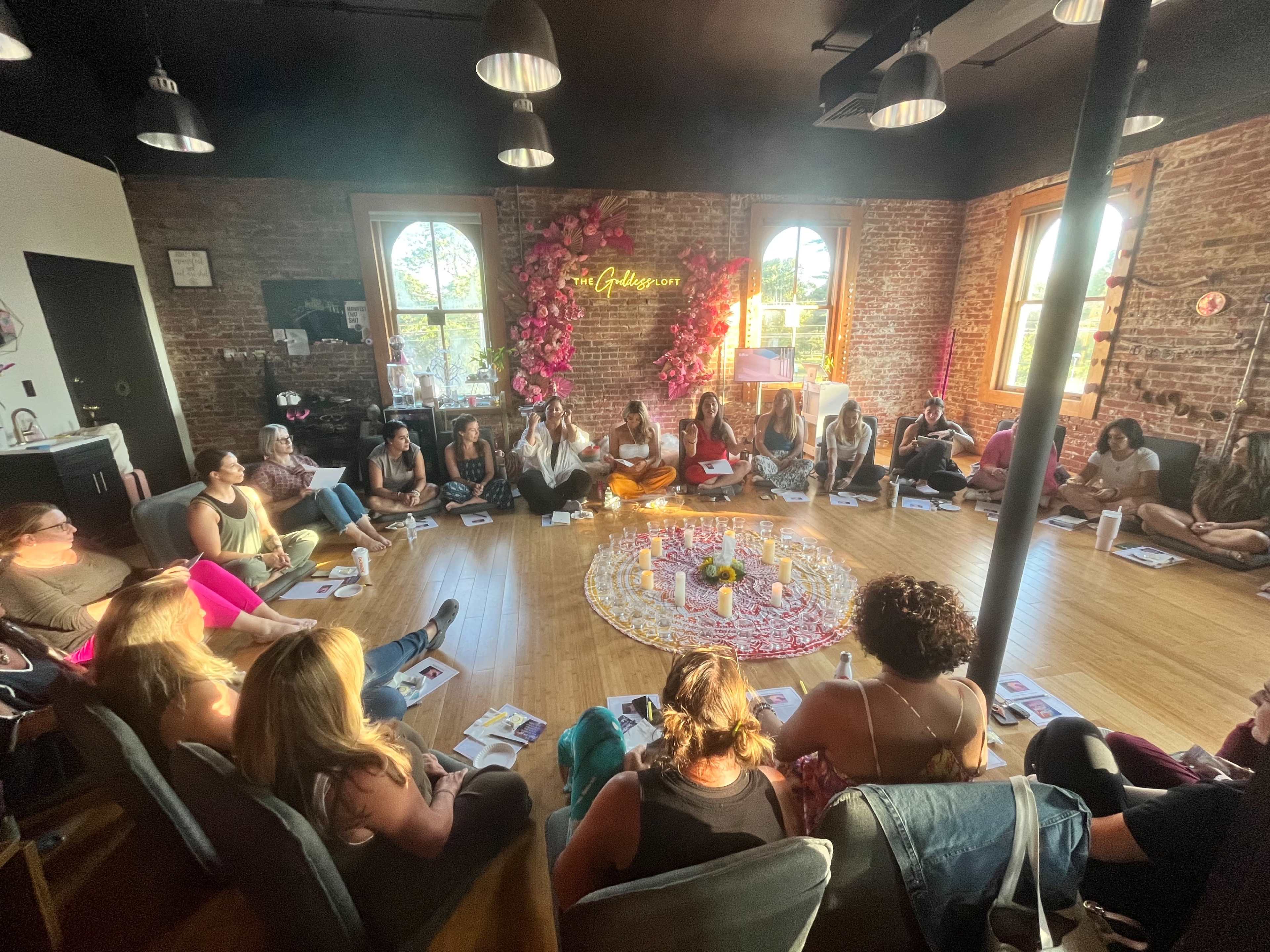 A group of people sits in a circle on the floor of a room with brick walls and large windows, surrounded by candles and a flower arrangement.