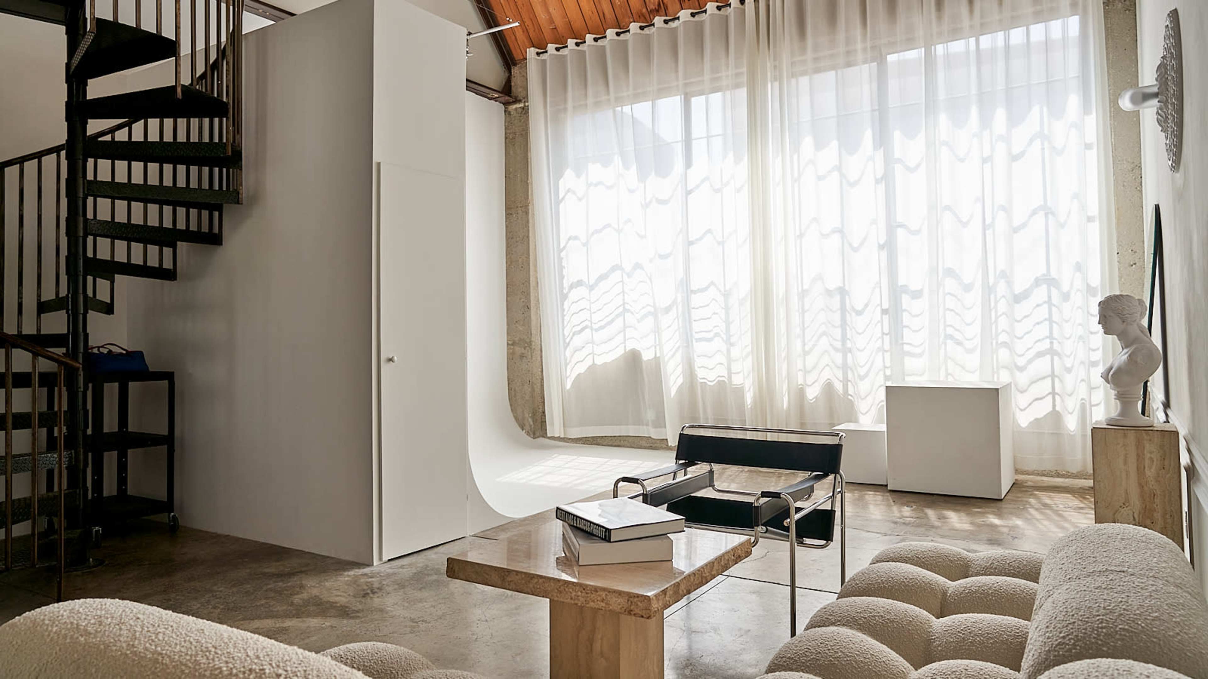 A modern interior space with a spiral staircase, large windows featuring sheer curtains, and minimalist furniture arranged around a central table.