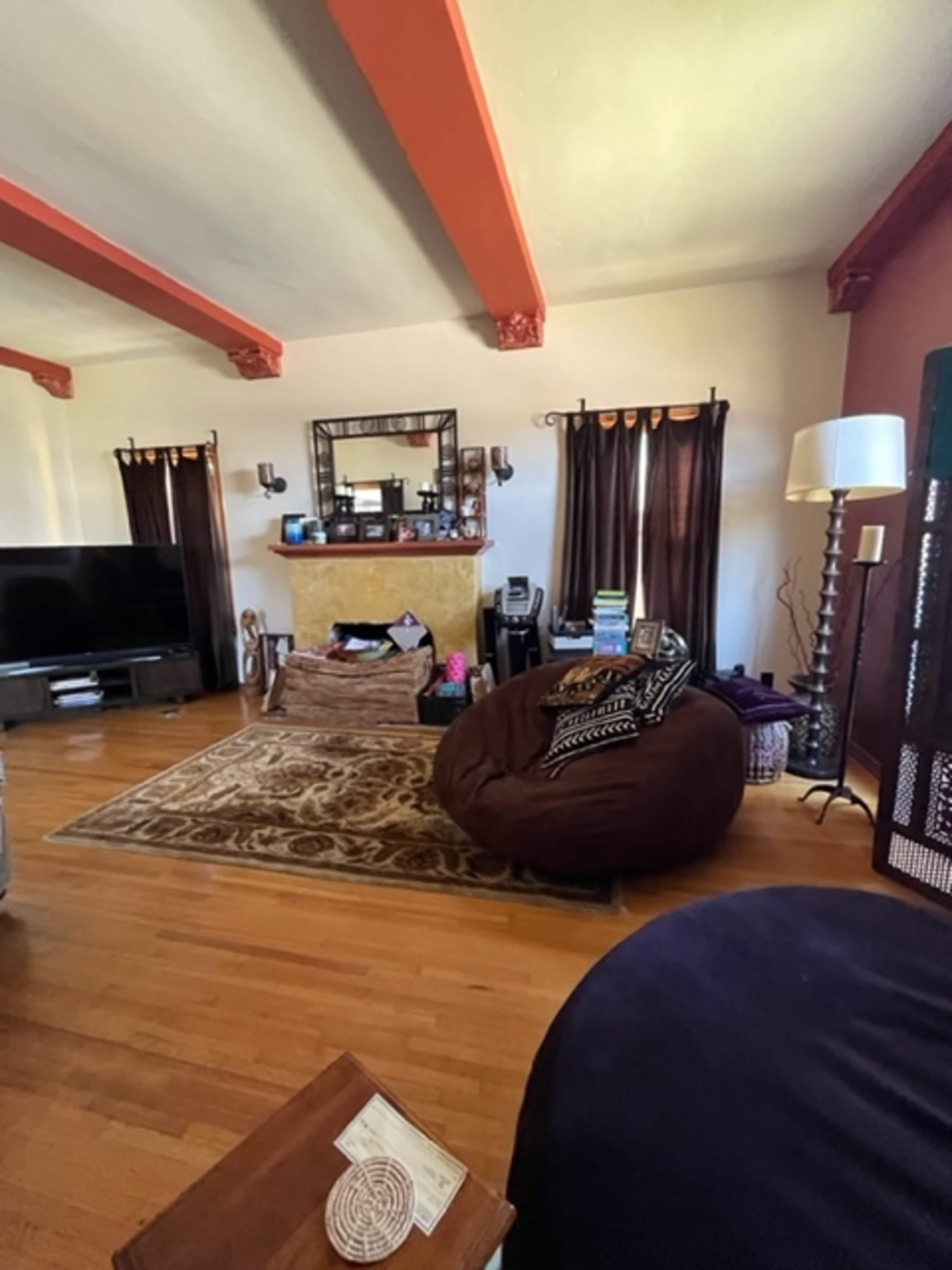 The image shows a living room with wooden beams on the ceiling, a large television, a decorative brown and gold area rug, and a cozy bean bag chair.
