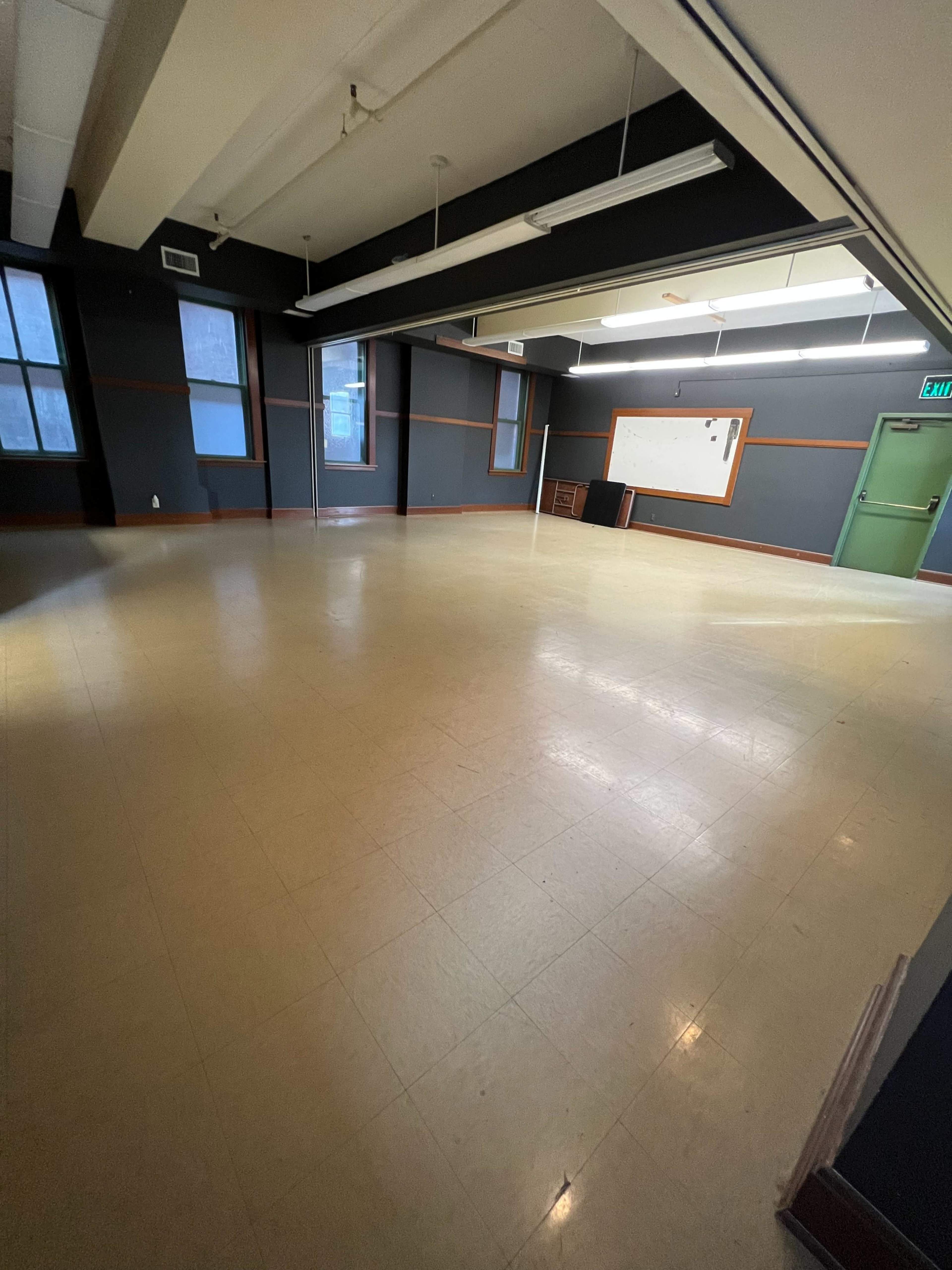 The image shows a spacious, empty room with wooden floors, large windows, and a chalkboard on the wall.