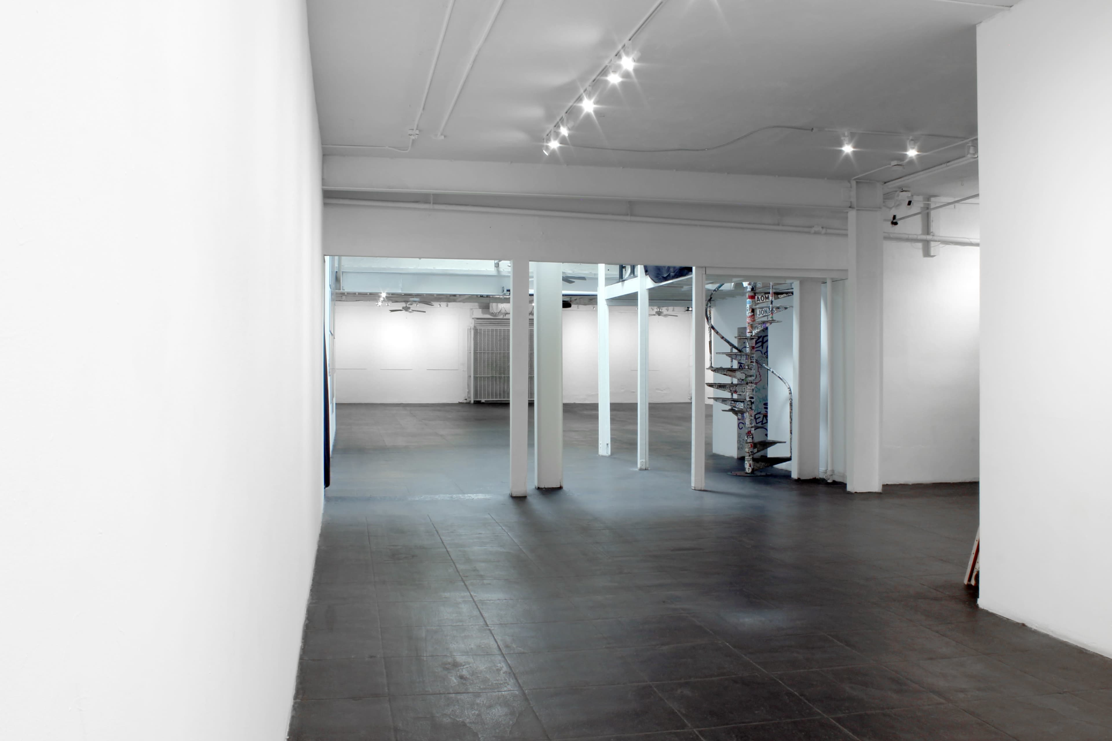 The image shows a spacious, minimalist gallery interior with white walls, a spiral staircase, and polished concrete floors.