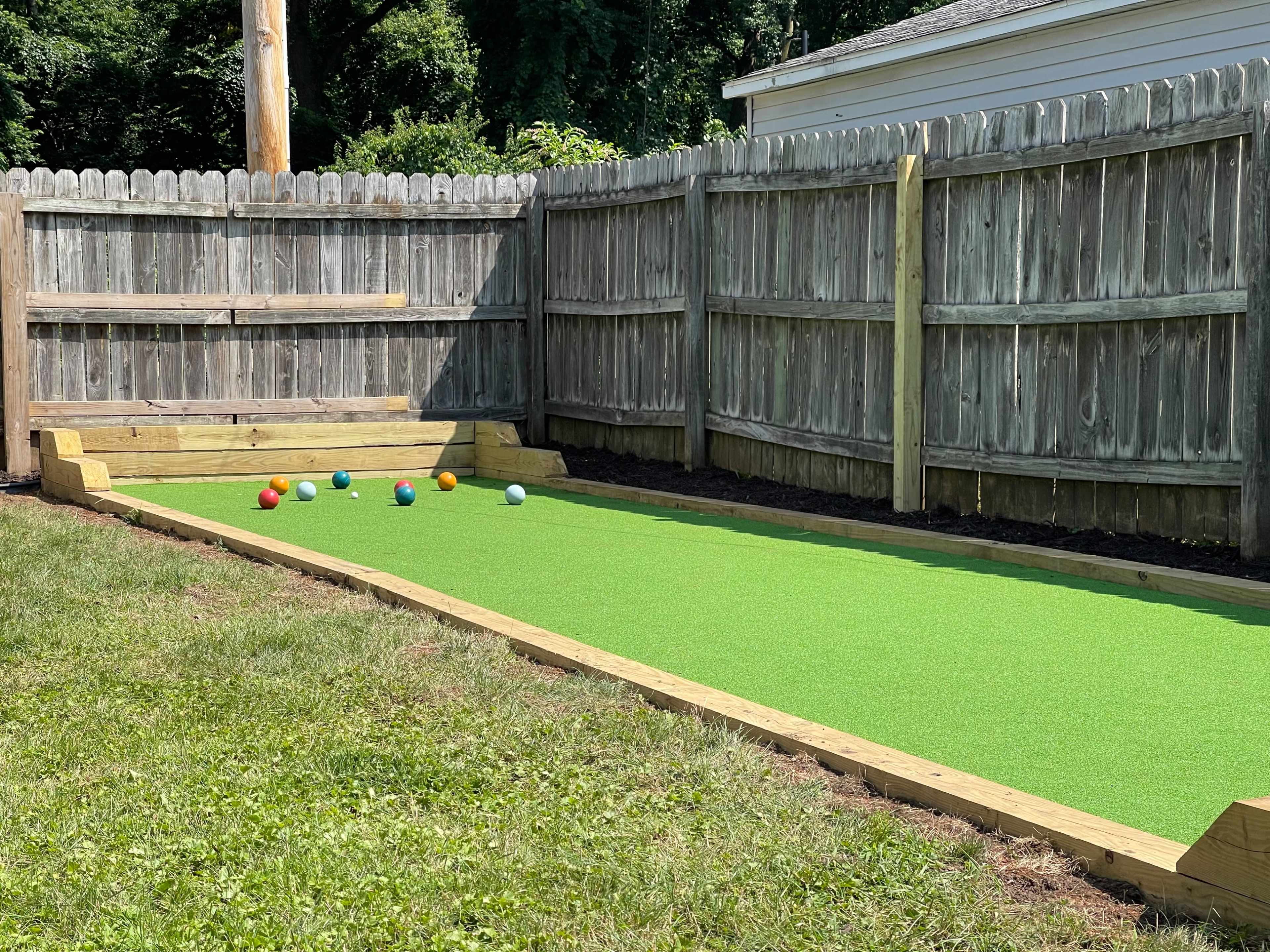 A synthetic green bocce court is located in a fenced backyard, featuring wooden borders and several colored balls arranged on the playing surface.