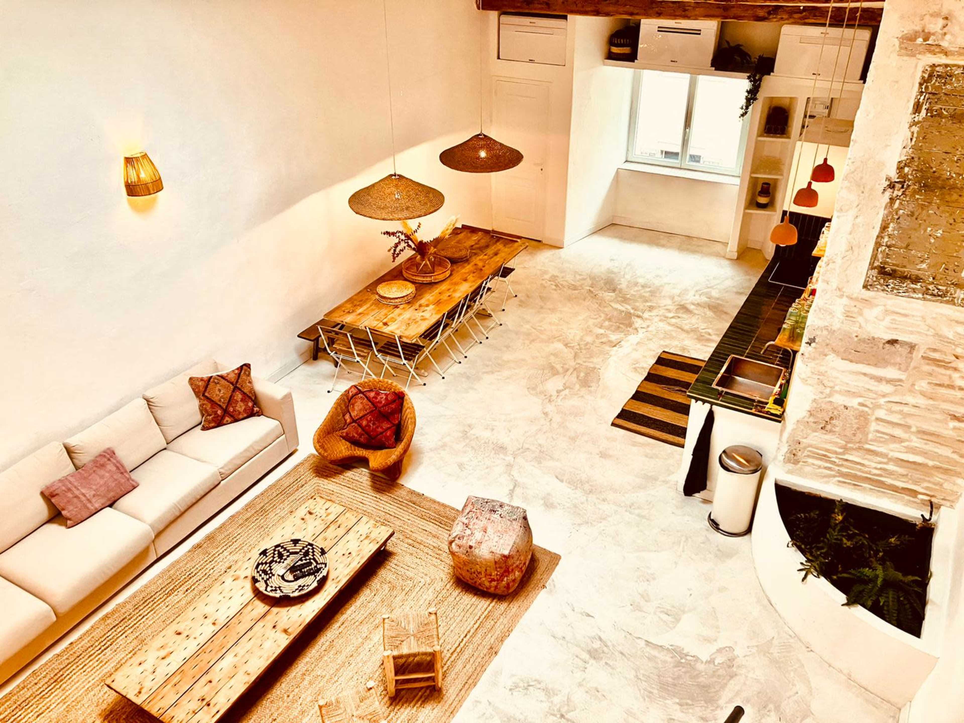 The image shows a spacious, modern interior with a large wooden dining table, cozy seating arrangements, and various decorative elements, all illuminated by warm lighting.