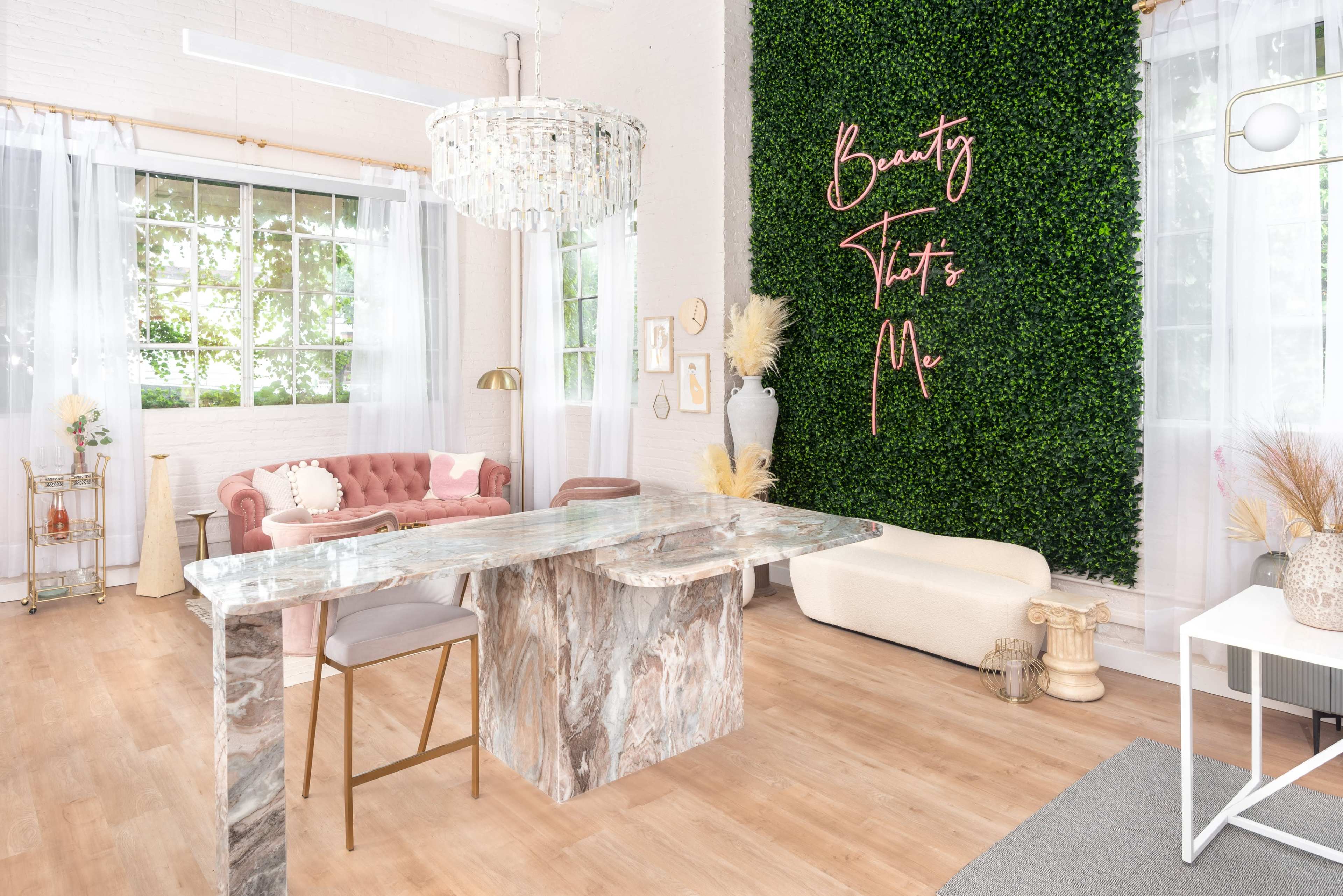 A modern interior space featuring a marble table, a pink velvet sofa, and a greenery wall with the words "Beauty That's Me" in elegant lettering.