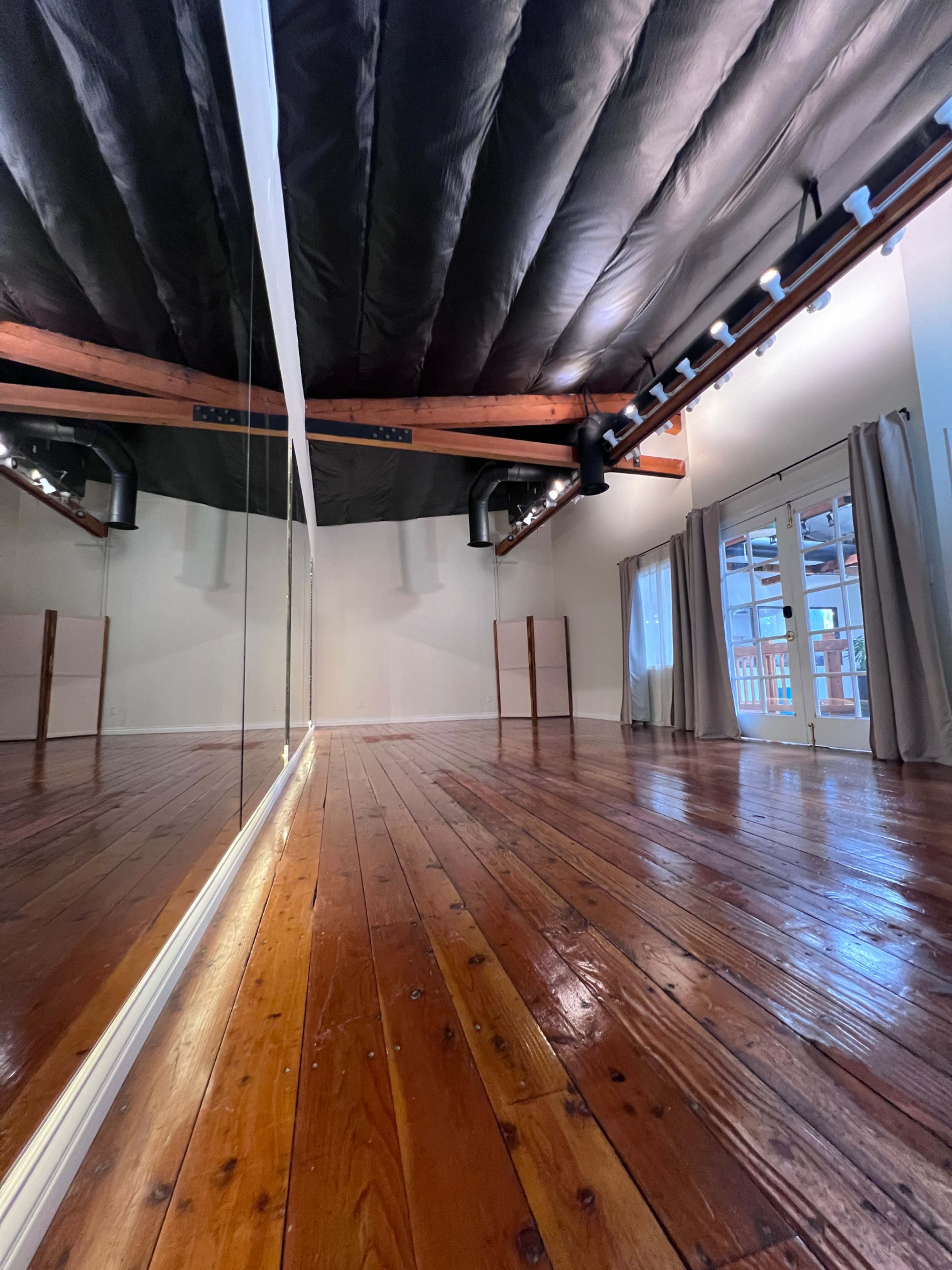 The image shows an empty studio with wooden floors, a large mirror on one wall, and windows with curtains providing natural light.