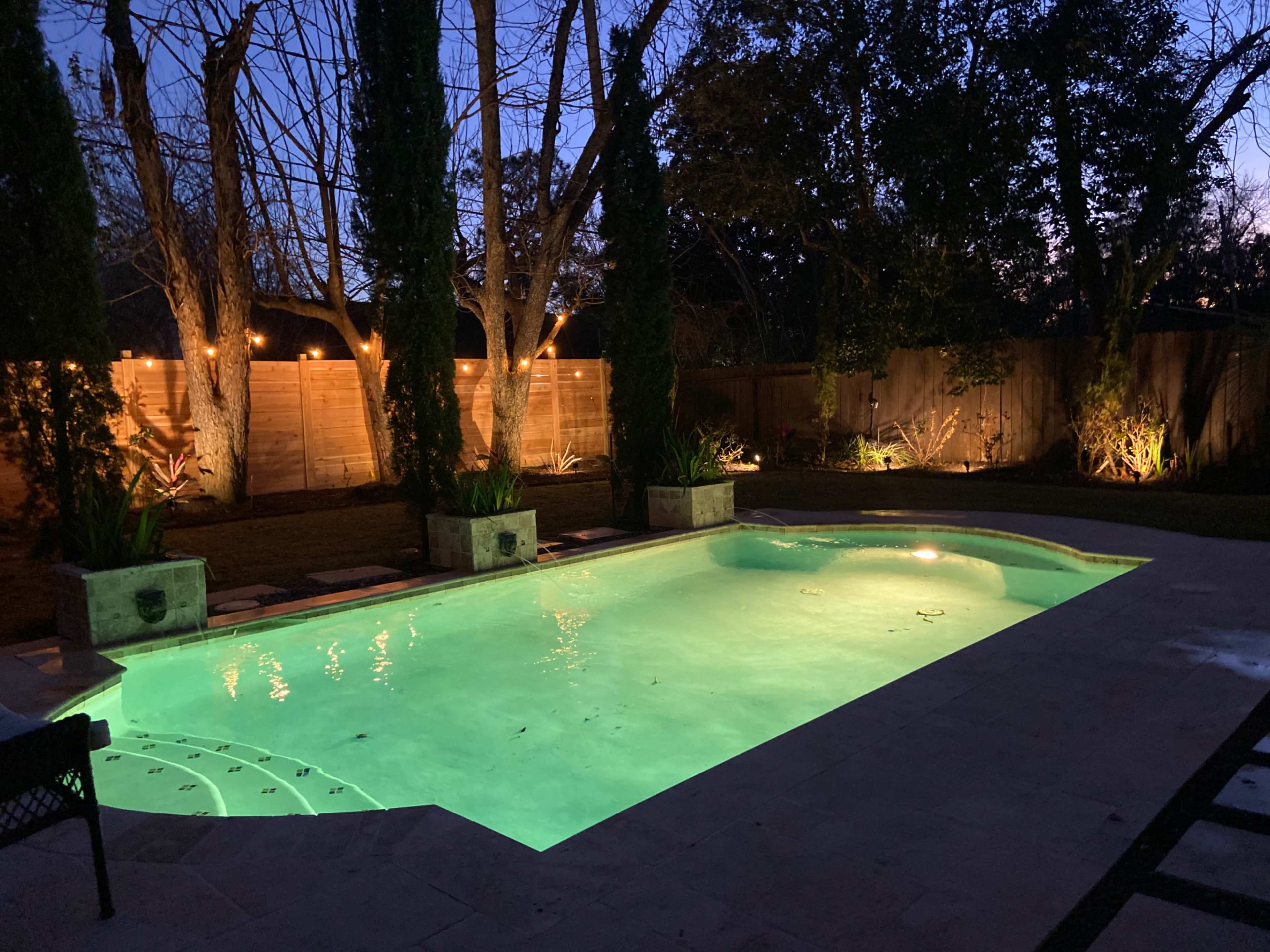 A well-lit swimming pool surrounded by landscaped trees and a wooden fence, with lights illuminating the area at dusk.