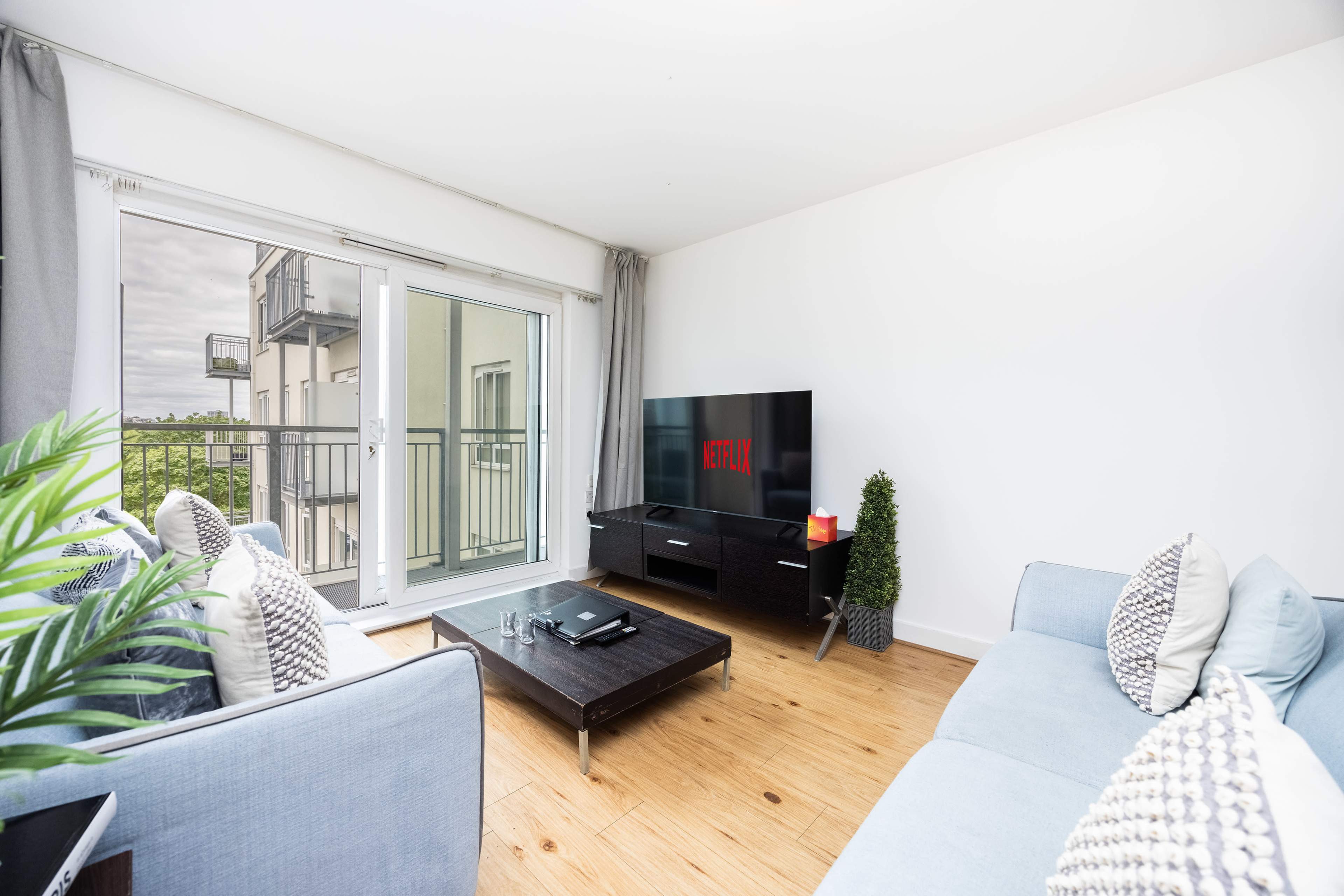 A modern living room features a light blue sofa, a coffee table, and a television displaying the Netflix logo, with large windows showing a view of a balcony.