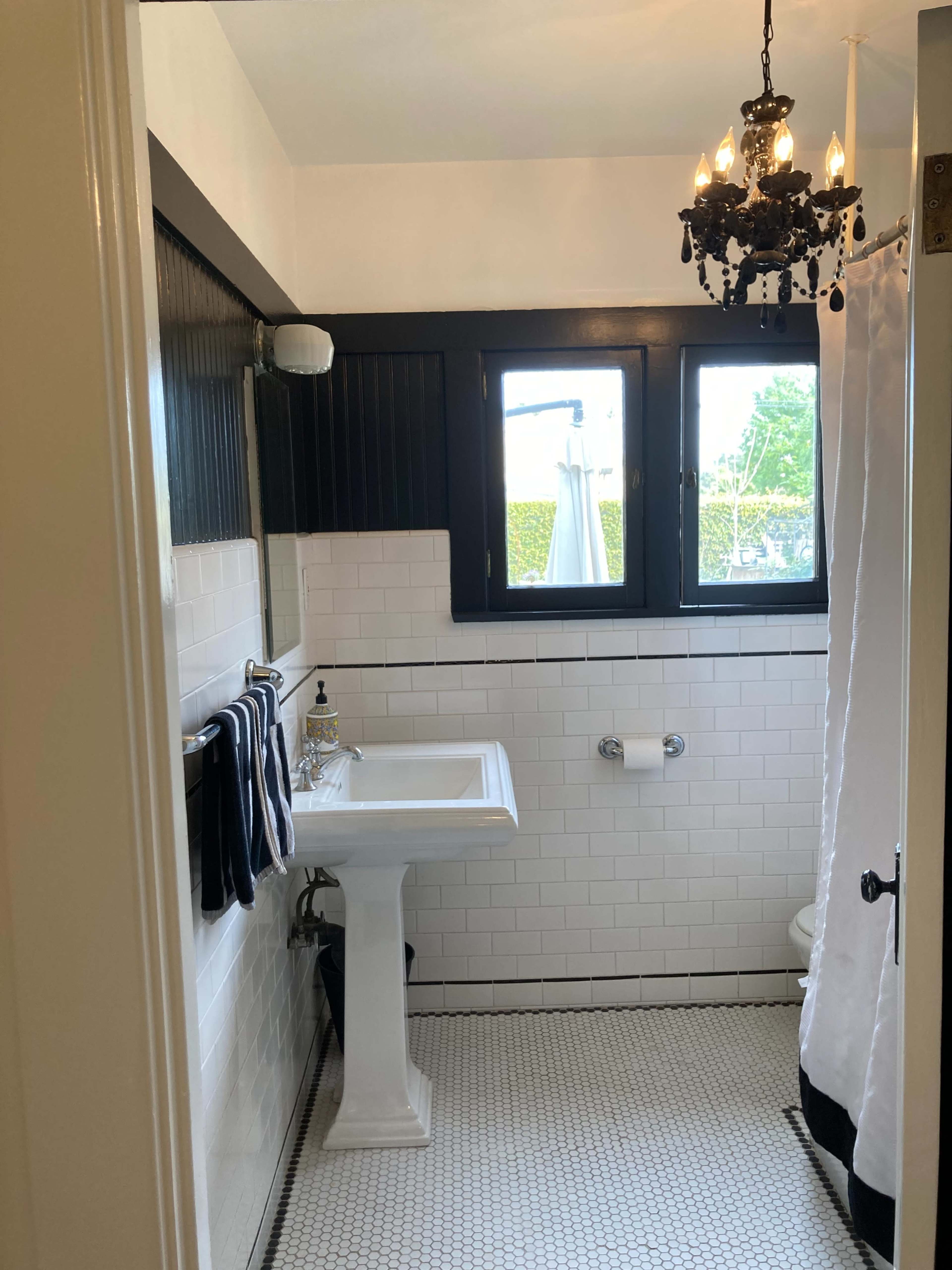 The bathroom features a pedestal sink, a shower curtain, and a vintage chandelier, all surrounded by black and white tile.