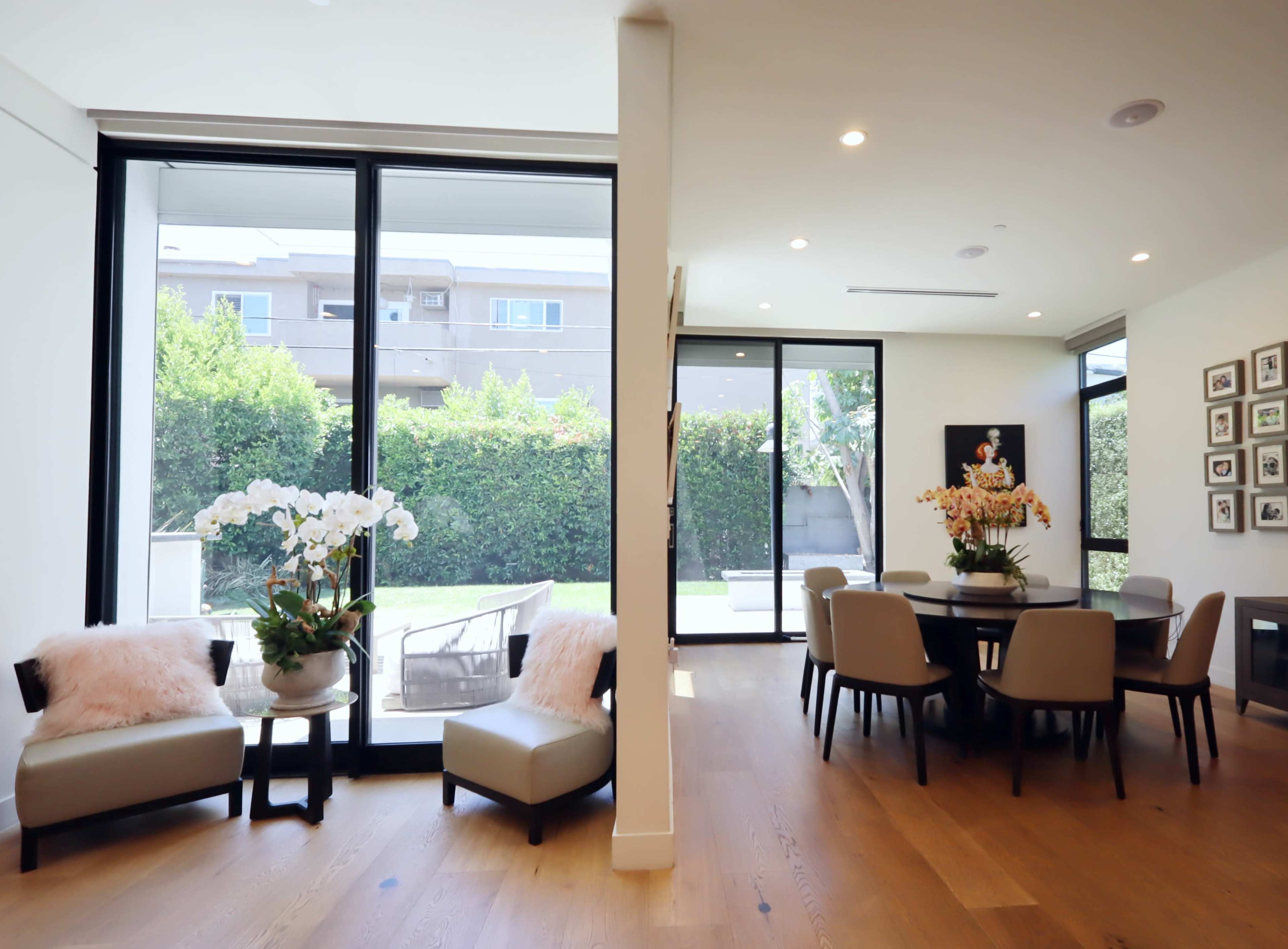The scene shows a modern dining area with a circular table, surrounded by chairs, adjacent to a lounging space featuring two small chairs and potted orchids, with large windows that open to a backyard view.