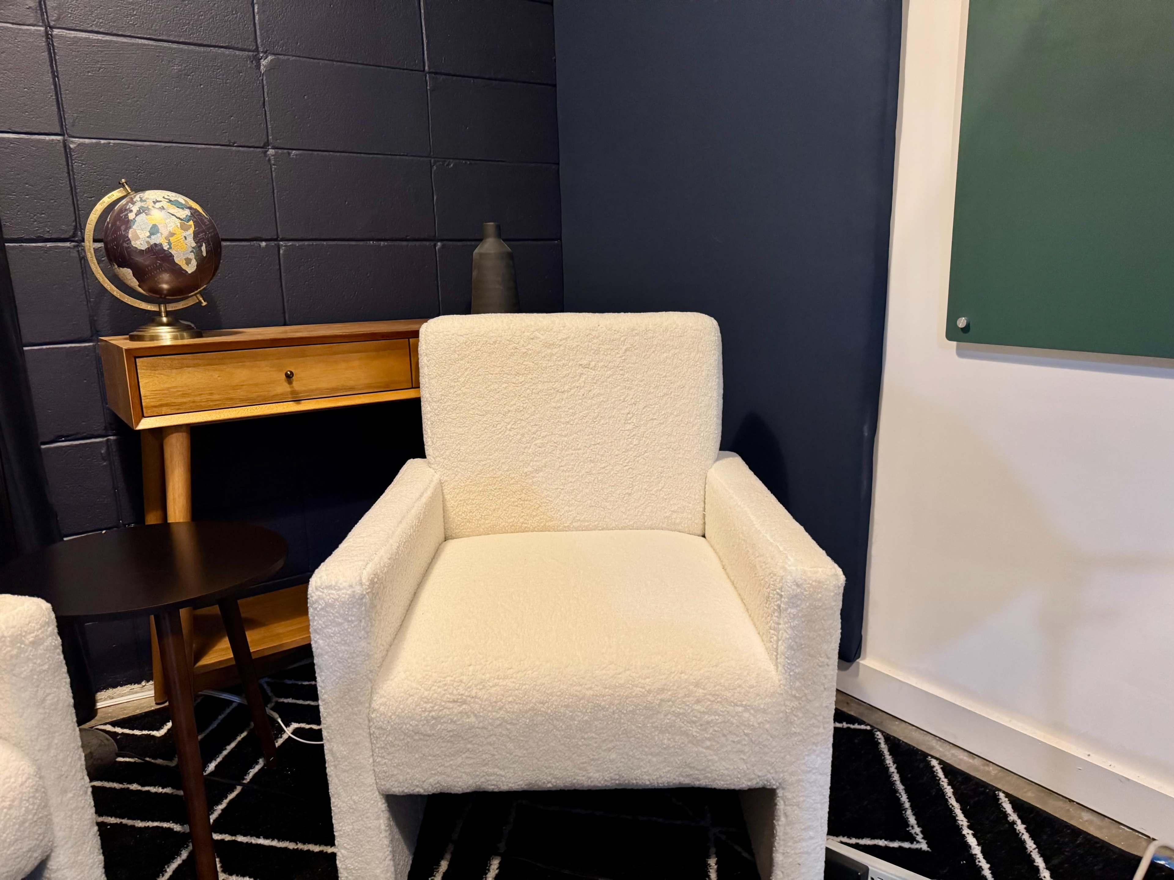 The image shows a cozy corner of a room featuring a white armchair, a small round table, a wooden side table with a globe, and a chalkboard on the wall.