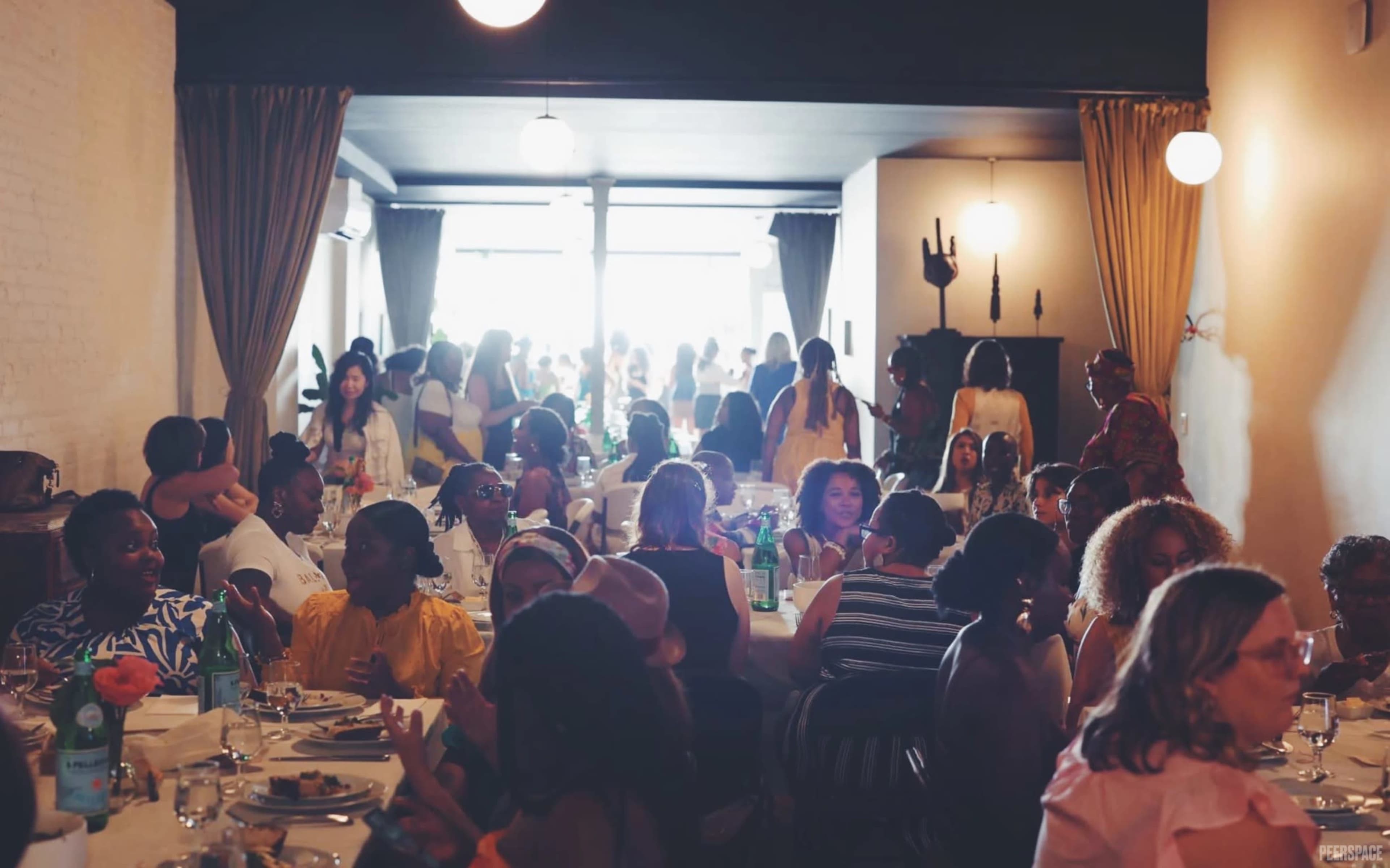 A group of women gathers around tables in a well-lit dining space, engaging in conversation and enjoying their meals.