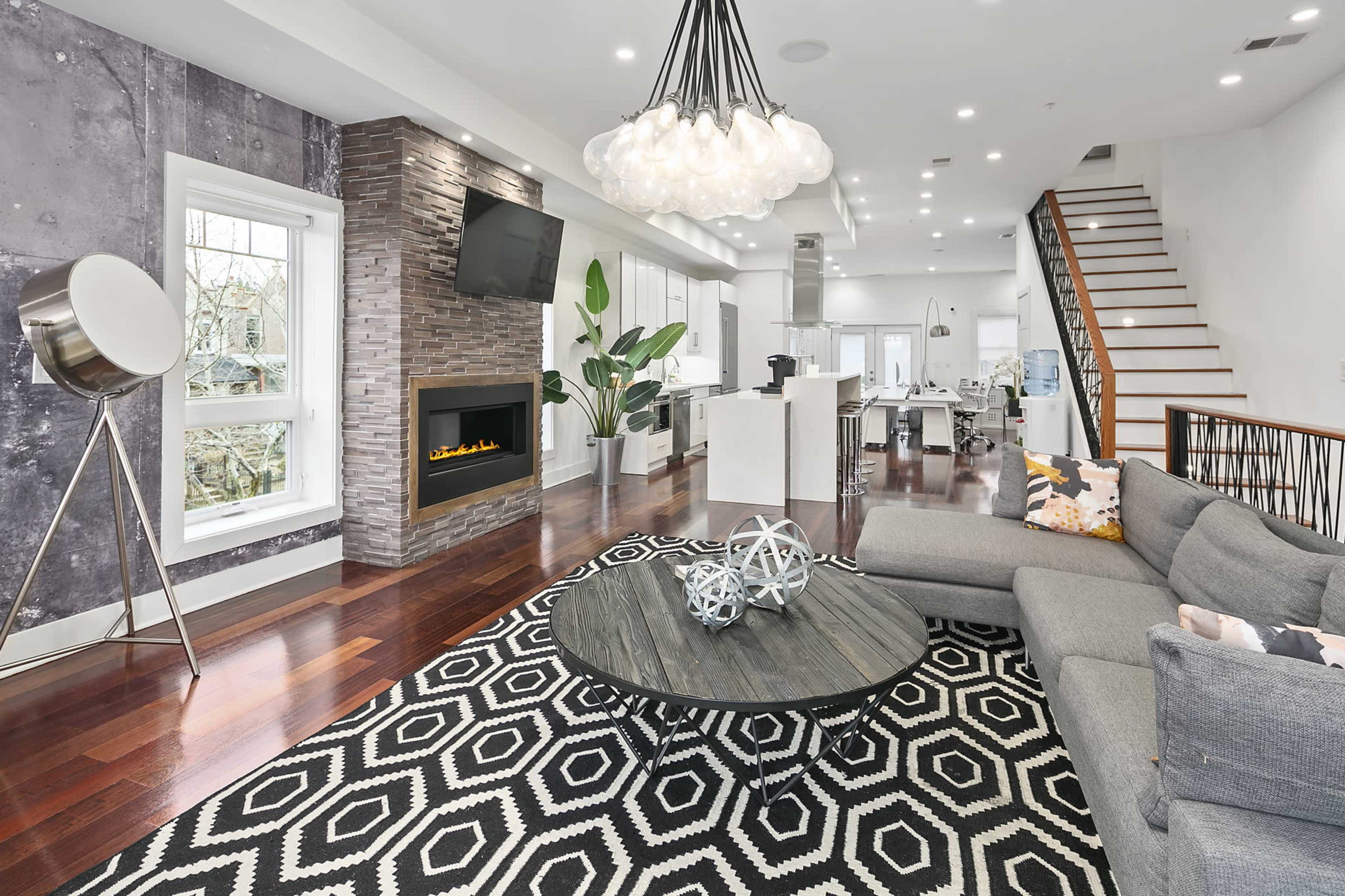 A modern living room with a gray sectional sofa, a round coffee table on a patterned rug, a wall-mounted television, a contemporary light fixture, and an electric fireplace.